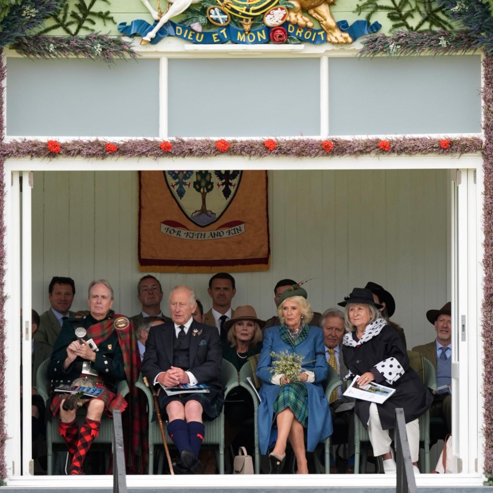 The King and Queen sitting with others in an outdoor viewing box or pavilion. King Charles is wearing traditional Scottish attire, including a kilt, as is the man next to him. Above the group is a decorative crest with a lion and the motto "DIEU ET MON DROIT". Beneath the crest is a banner reading "FOR FAITH AND KING".
The pavilion is decorated with greenery and red flowers along its top edge.