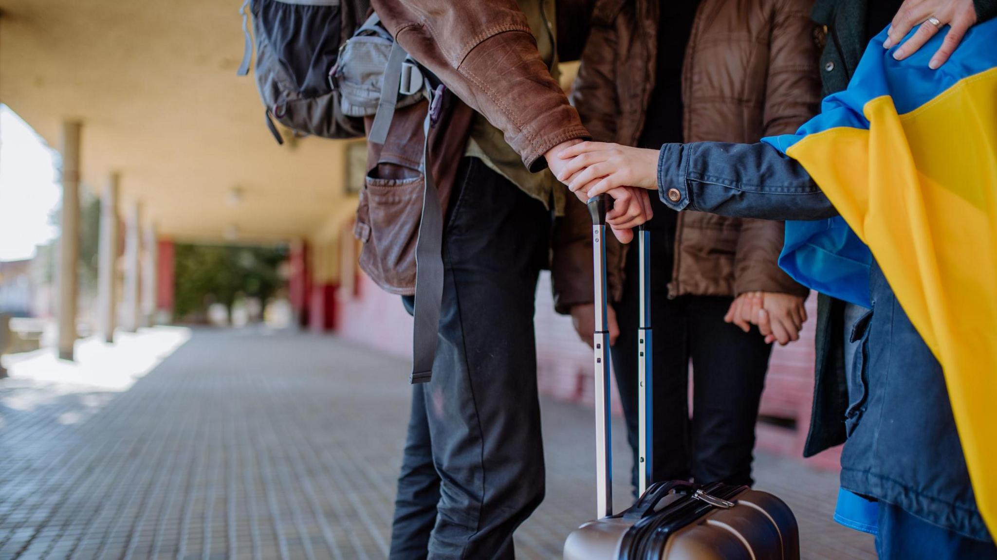 A mid-level shot of three people standing on a concourse. One is a child in a blue jacket with a Ukrainian flag draped across their shoulders. They are holding hands with an adult in a brown jacket. Another adult in a brown jacket has their hand on a suitcase.