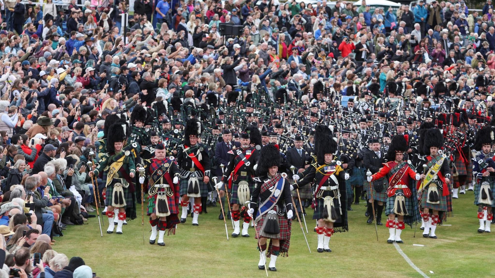 A marching band in traditional Scottish attire. Band members are wearing kilts and feathered hats, and are playing bagpipes and drums. Some are carrying batons. A large crowd surrounds them, with people both seated and standing, watching the performance attentively.
