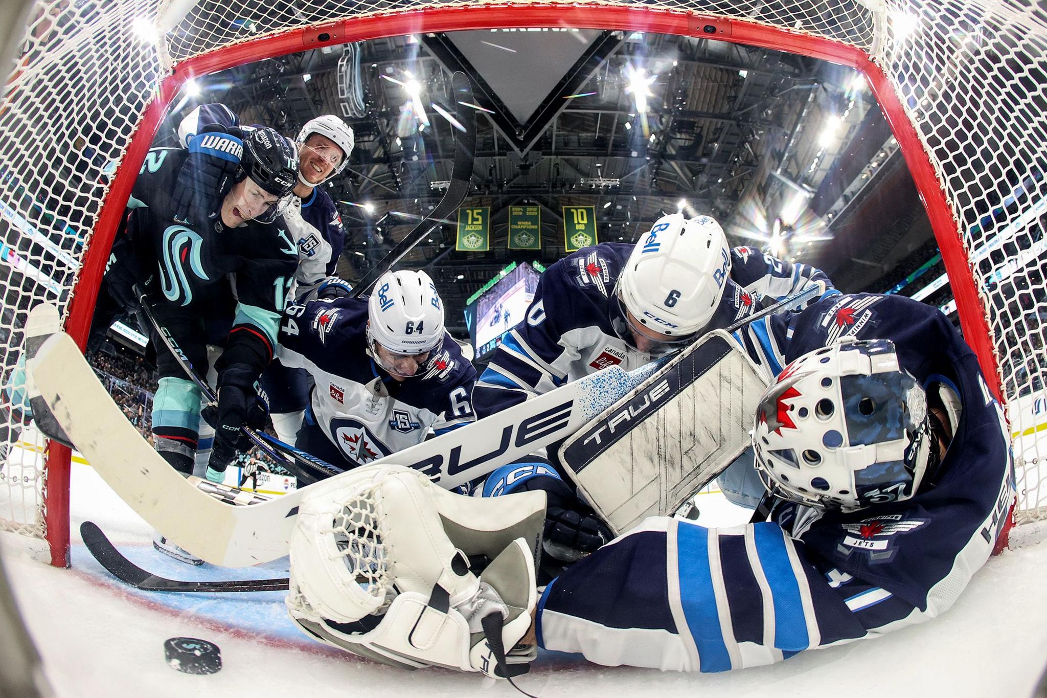 Ice hockey players battling for the puck inside the goal crease, with the goalie in blue gear sprawled on the ice and teammates crowding around under bright arena lights.