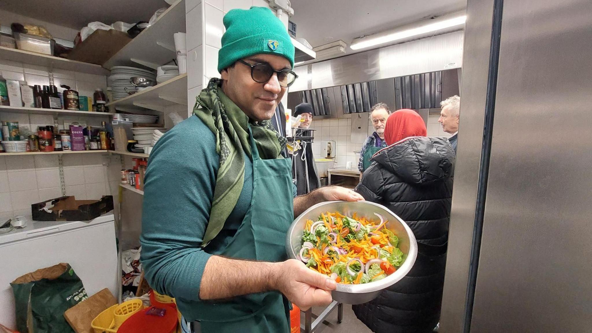 Sâmân Jamshidifard in a kitchen, holding a bowl with a vegetable salad in it. On the shelves behind him, there is cutlery, crockery and various cooking ingredients. Three men and a woman are also in the kitchen, having a conversation behind him.