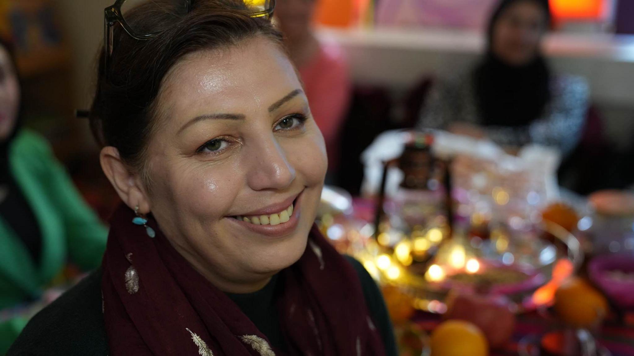 Rona Panjsheri has her hair in a ponytail and is smiling at the camera.
She is wearing turquoise drop earrings and glasses on her head. In the background are other women sitting around a table. 