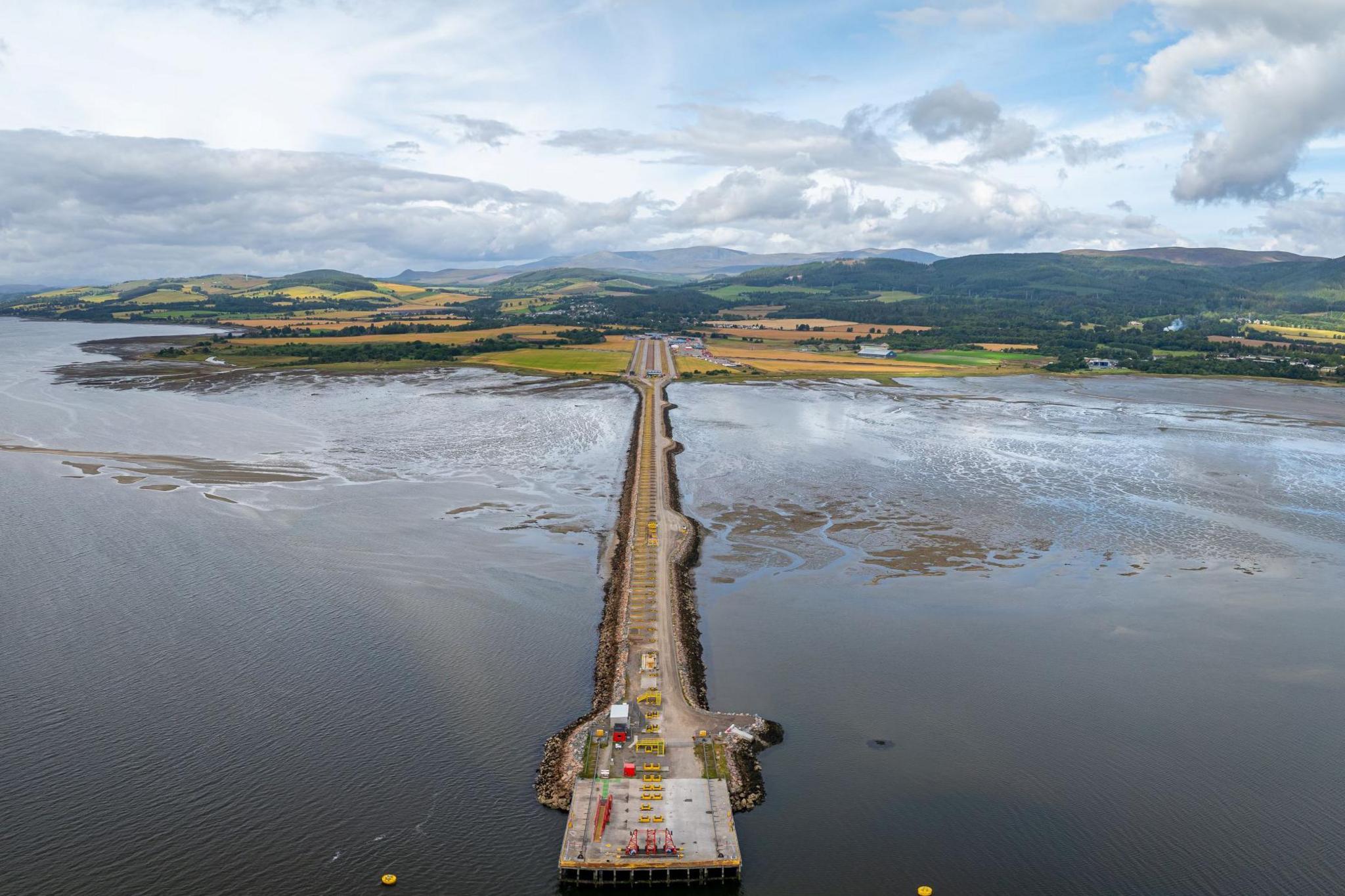 Highland Deephaven is on the shores of the Cromarty Firth. There is a long causeway extending out into the firth and ending in a square area of quayside. In the distance is a rural landscape of farms and fields.