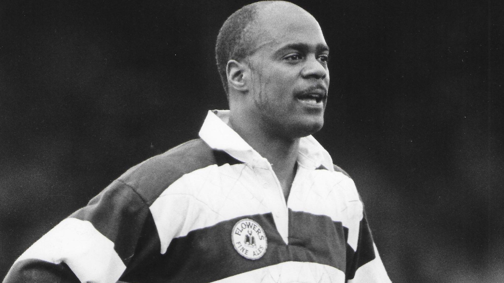 Black and white photo of Ralph Knibbs. He is pictured looking away from the camera at a rugby game. He is wearing a striped polo shirt with long sleeves. He has short hair and facial hair on his upper lip. His hands are on his hips. 