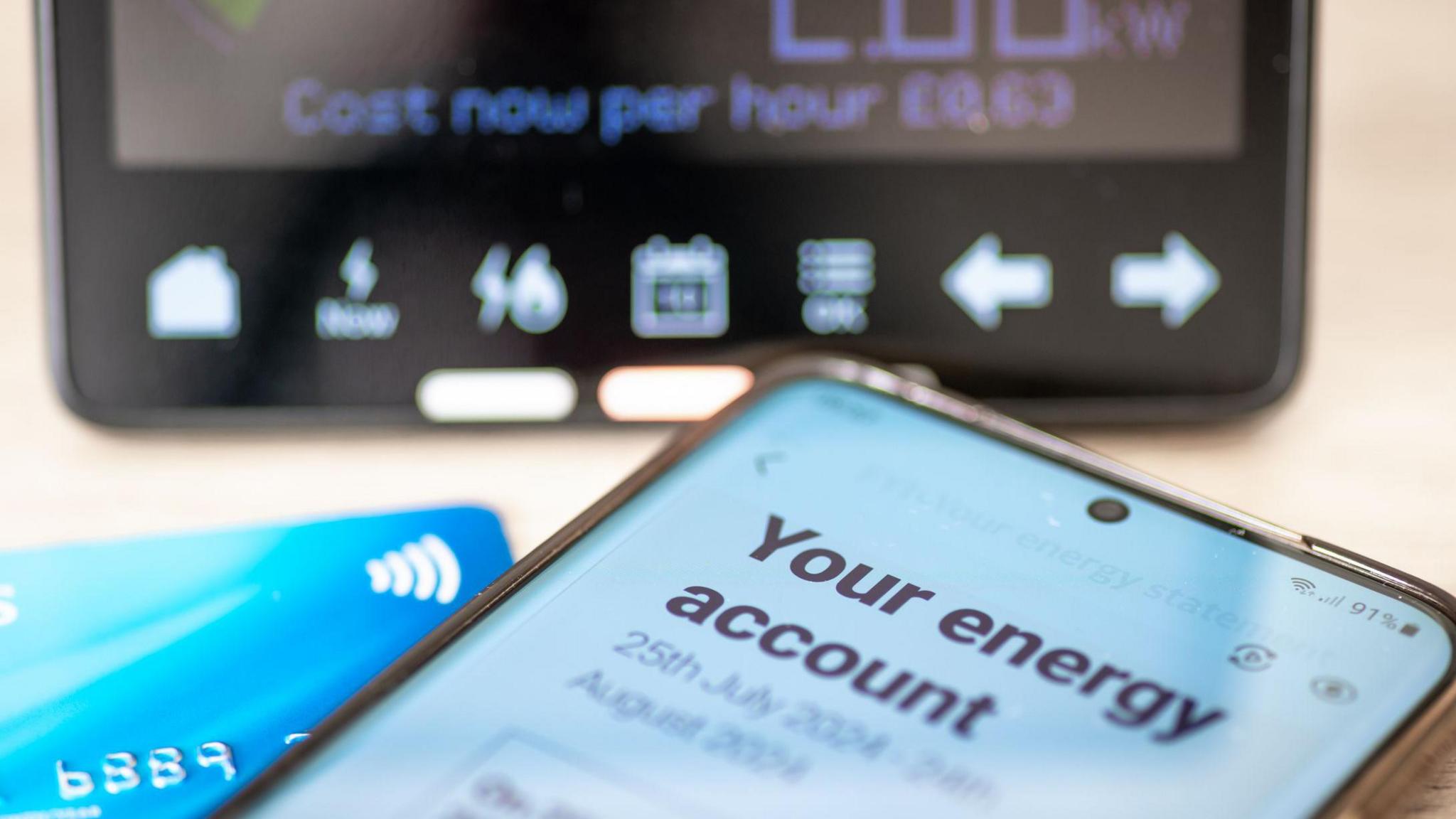 Smartphone with a page open on somebody's energy account, sits next to a debit card and in front of a smart meter display unit. 