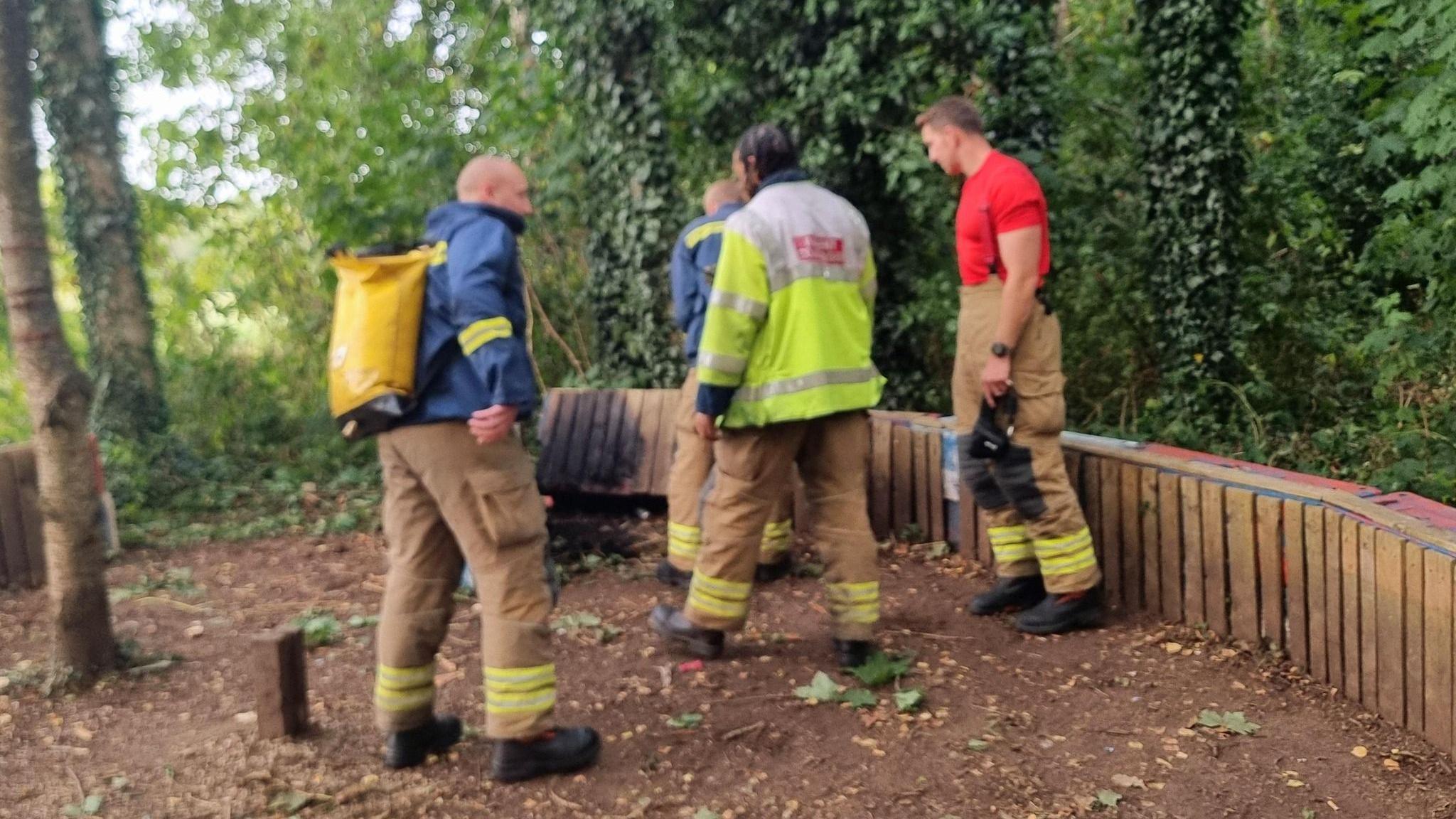 Four firefighters stand in a wooded area where a wooden board can be seen, charred from a fire