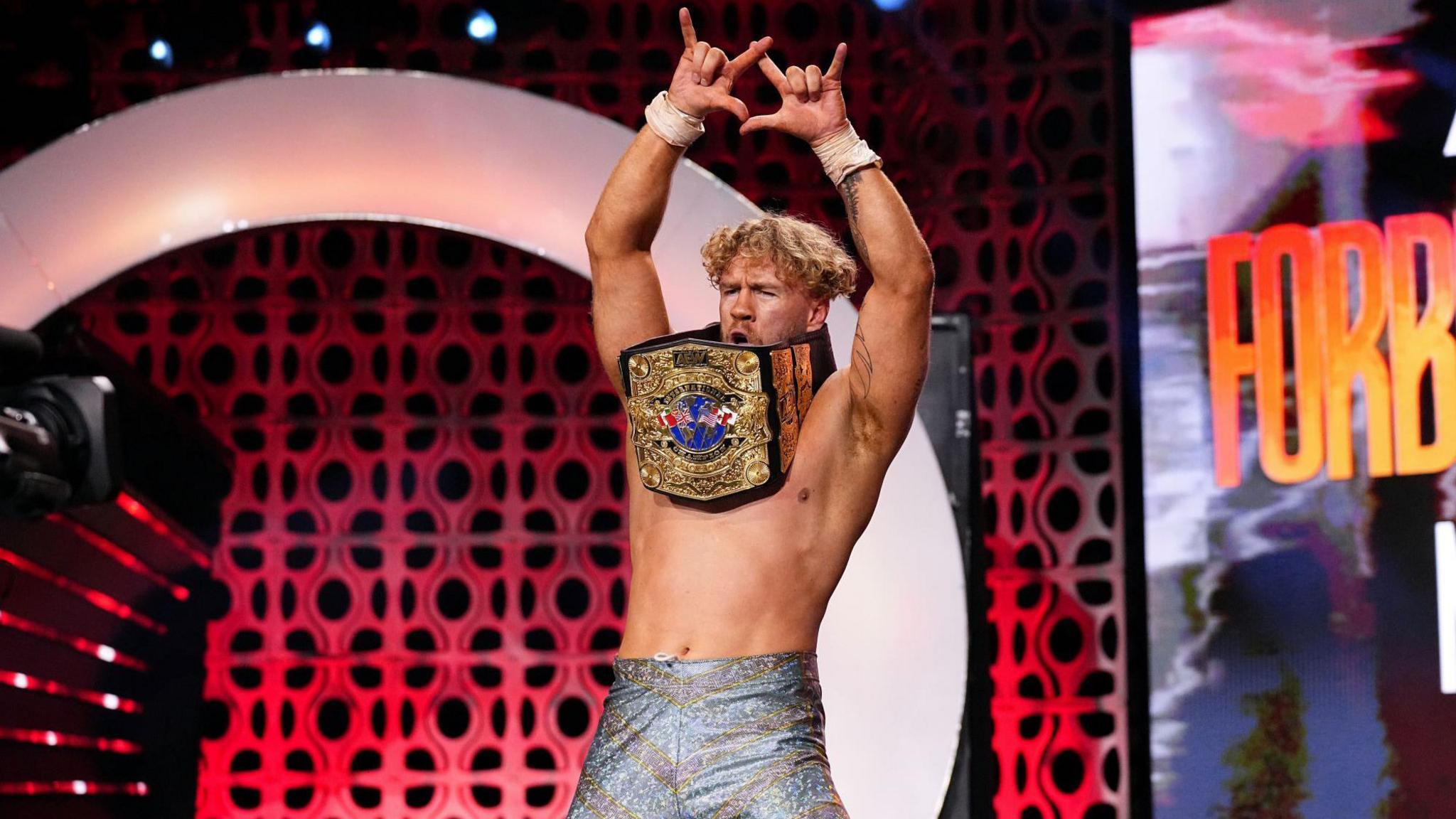A male wrestler standing with both arms above his head, making a 'devil horn' sign with both hands, with a wrestling belt draped around his neck. He has strawberry blond curly hair, is wearing tight sparkly trousers and is topless. 
