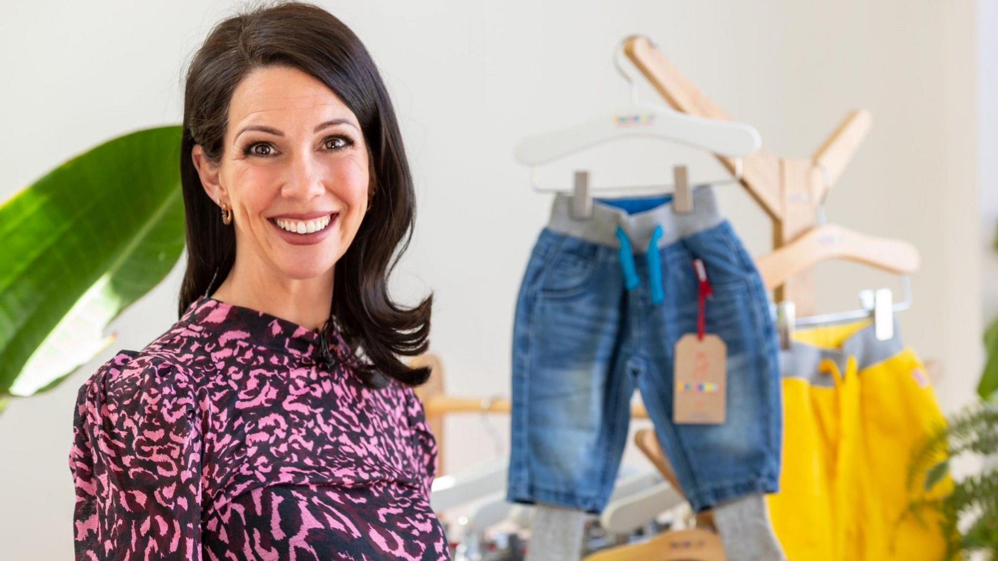 Natalie Hull wearing a pink and black top, smiling at the camera with a pair of blue denim sockatoos trousers in the background.