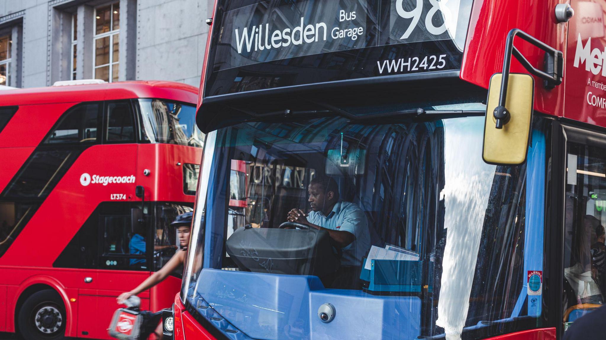 A bus driver sat in the front of a red double-decker London bus travelling to Willesden, leaning his arms on the steering wheel with his hands crossed. A cyclist is passing his bus on the left, and there is another red double-decker bus in the background.