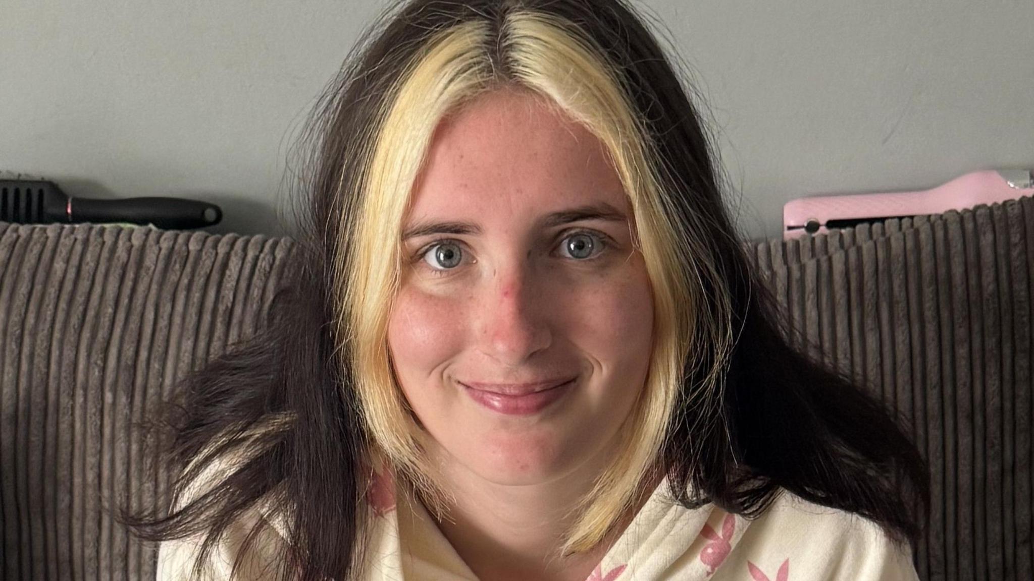 A close-up photo of Kailee who looks into the camera with a smile, she is sat on her sofa at home. She has dark hair down over her shoulders, apart from the front which is dyed blonde.
