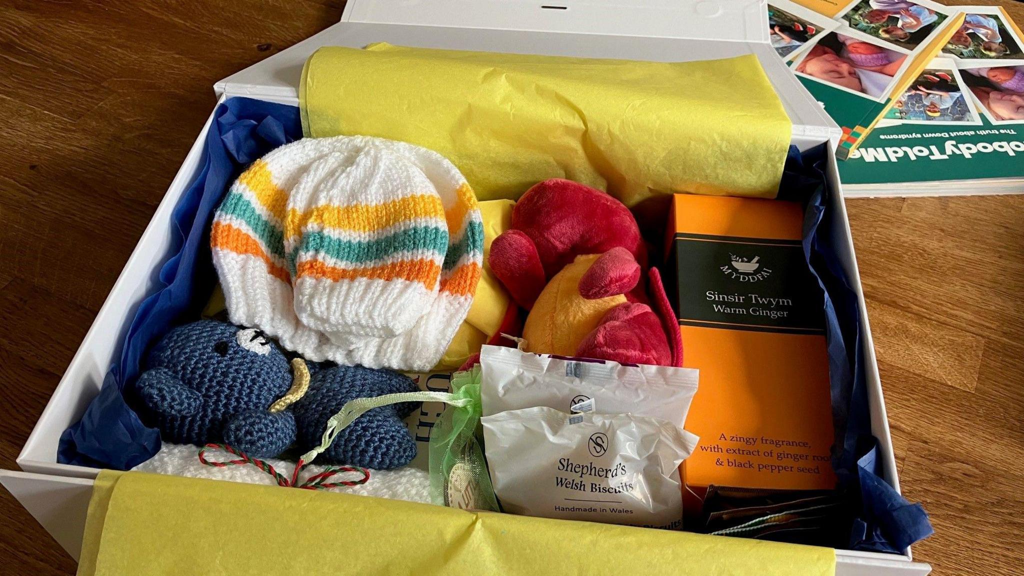 A cardboard box with blue and yellow tissue paper is filled with a knitted bear, a toy dragon, a woolly hat and some biscuits.   