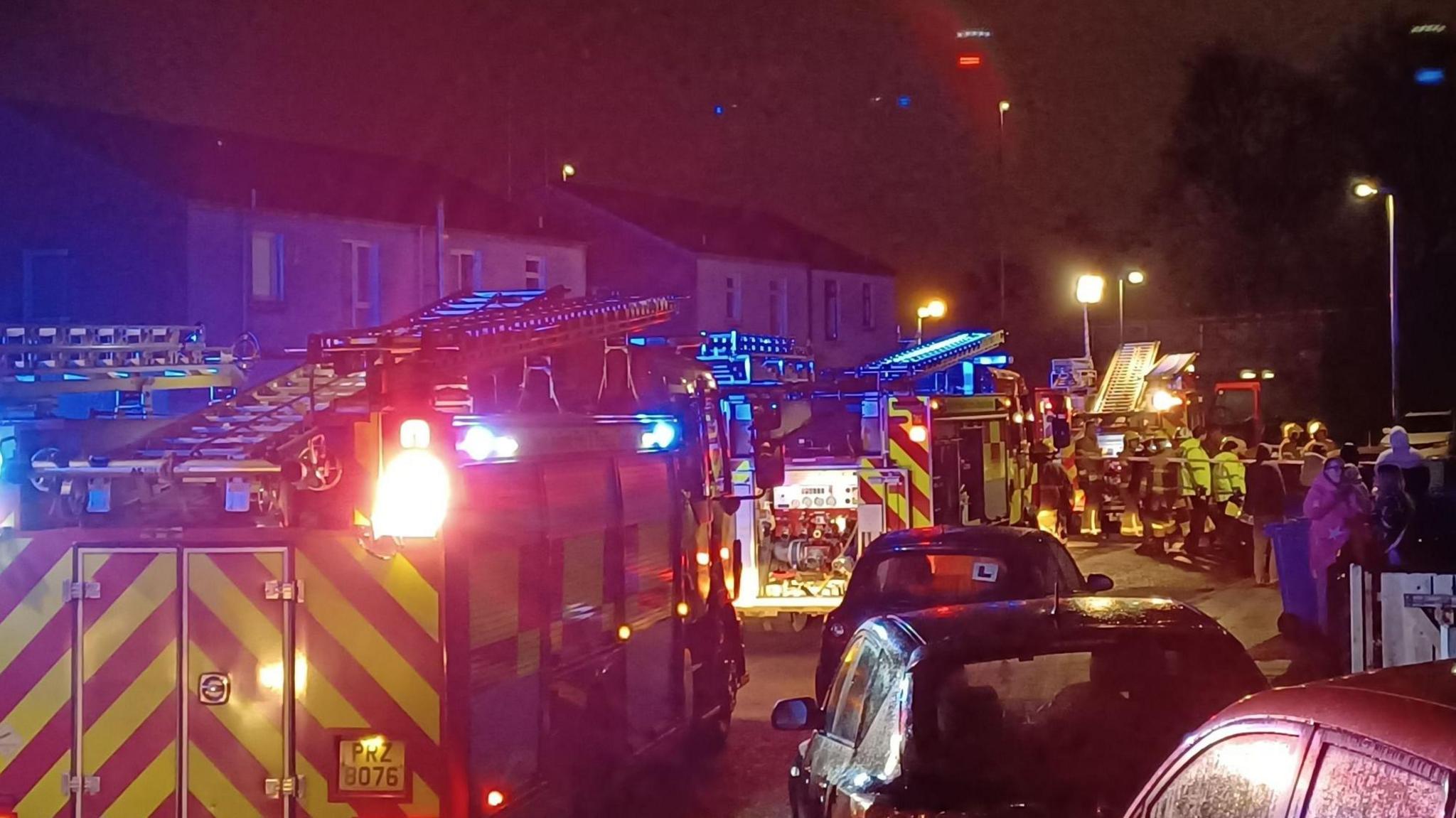 Fire engines, red and yellow in colour with flashing blue lights parked up on a street with parked cars around them. It's nighttime so the street lights are on and there are people standing on the sidewalk as firefighters in yellow uniforms and hard hats are moving equipment across the road. 