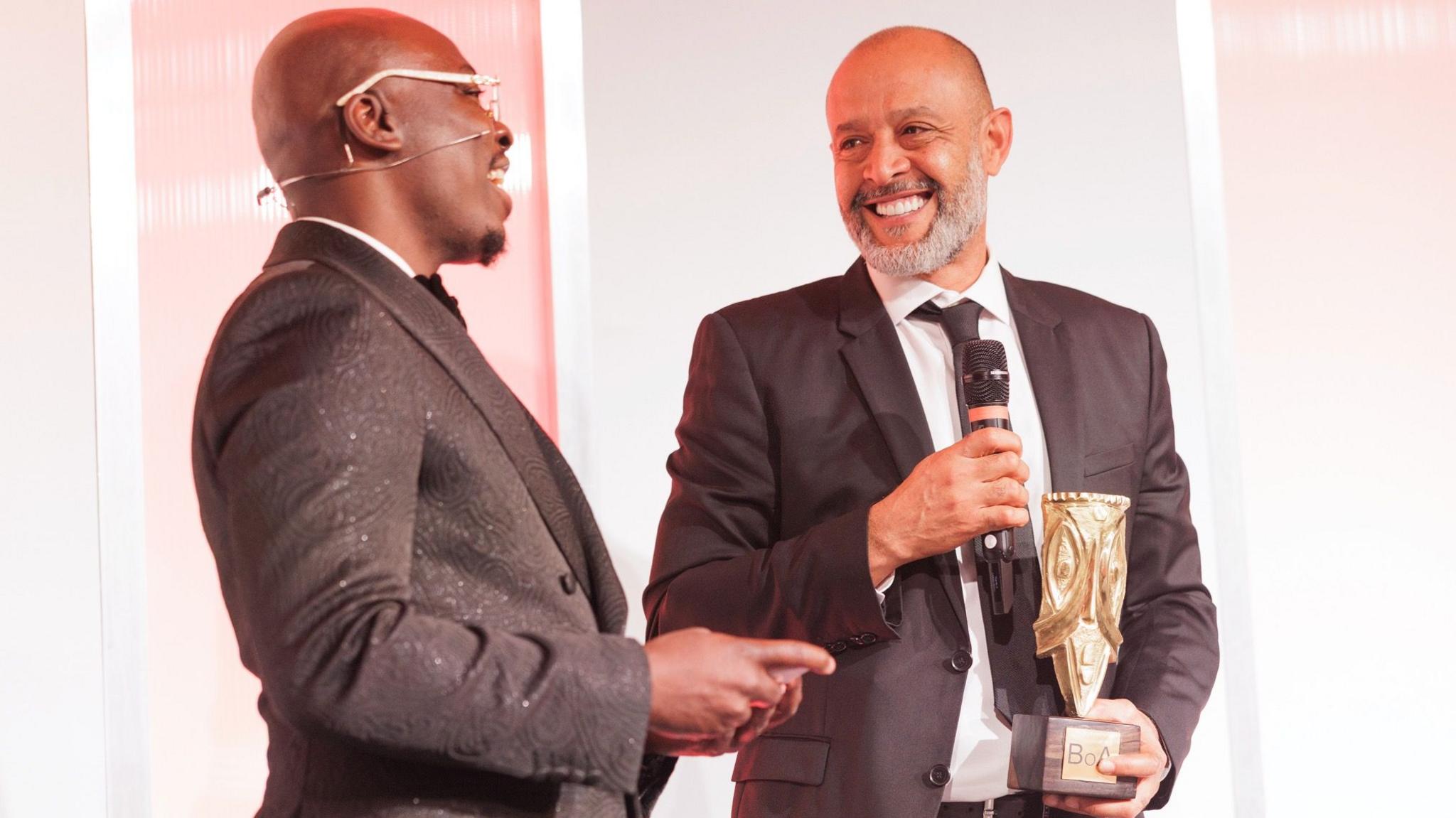 Edi Kadi and Nuno Espirito Santo are seen from waist up wearing suits during the 2025 Best of Africa Awards dinner. Kadi, who is also wearing glasses with white frames, is talking while Nuno is smiling and holding a black microphone in his right hand and a golden Best of Africa award in his left. Both men are turning their necks slightly to look at each other while on stage