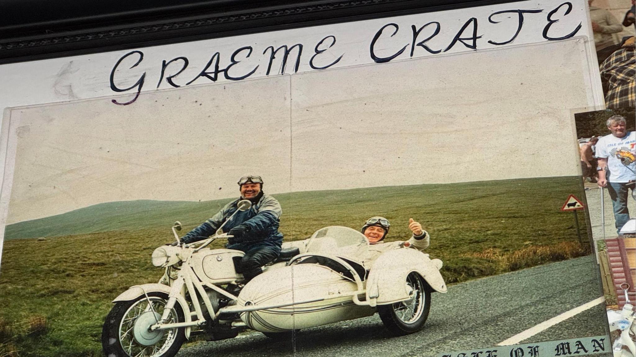 A photo of a display which has a photo of Graeme Crate riding his white BMW sidecar, he is on a road near a green hill. He wears a blue and grey coat, with goggles on his forehead, and has a passenger who wears goggles, a cream coat and is doing a thumbs up.