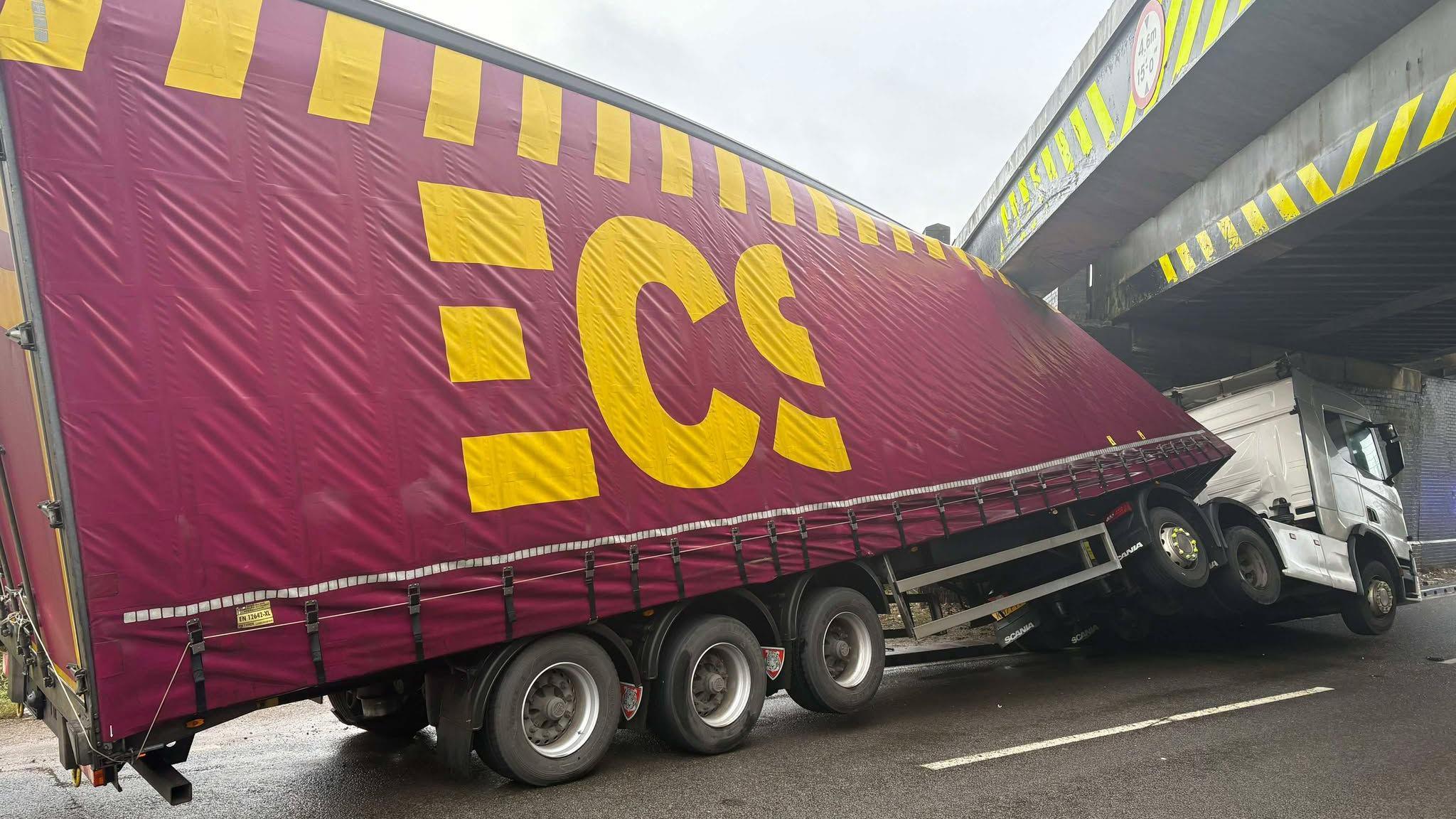 An image of a large lorry that has been involved in an accident underneath a bridge. The truck, which is maroon and yellow, is angled and appears to have been damaged on impact and displaced.