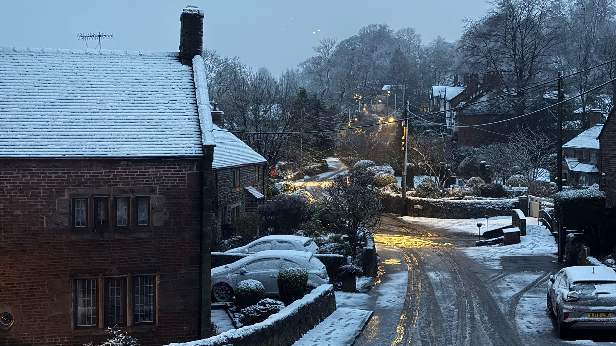 A snow-covered village with houses and cars on either side of a road, which is covered in melted snow. Trees can be seen in the background as the road curves to the left.