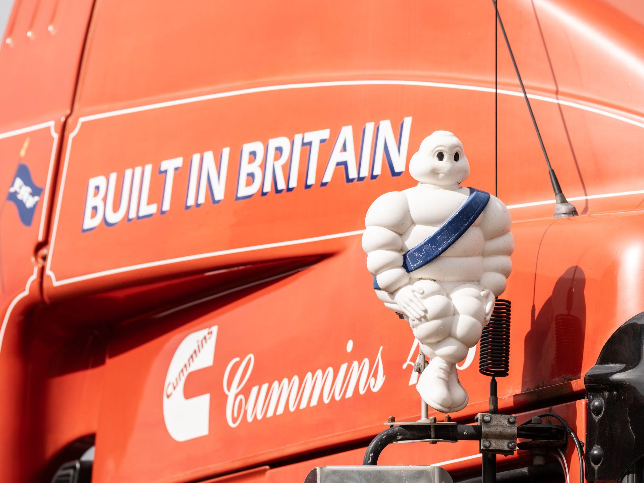 Classic red vehicle with Built in Britain in lettering on the side. A white Michelin Man figure is mounted on the vehicle. It is wearing a blue sash. 