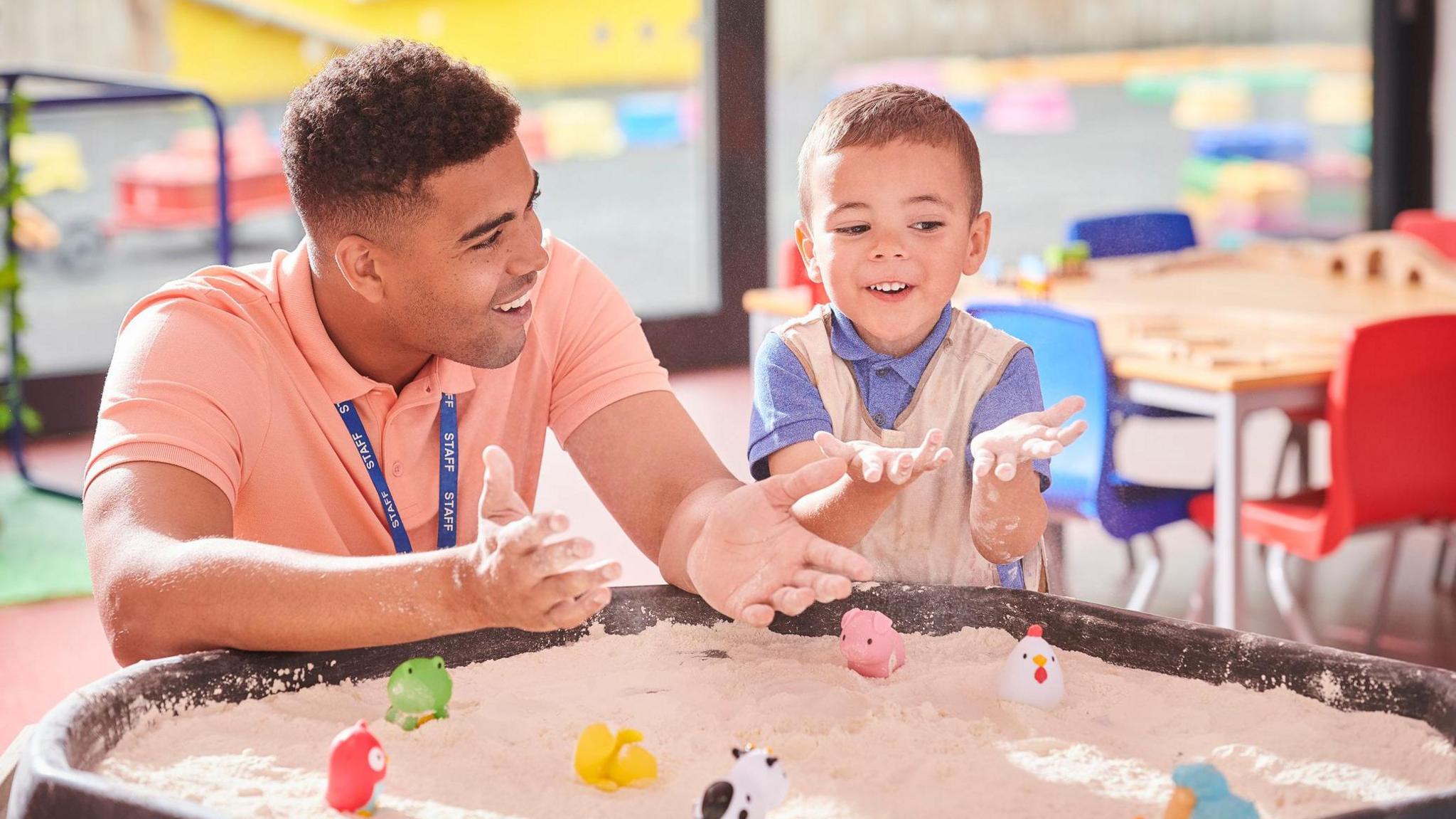 A nursery worker wearing a blue lanyard reading "staff" and a small boy wearing an apron play with a sandpit full of plastic animals at nursery.