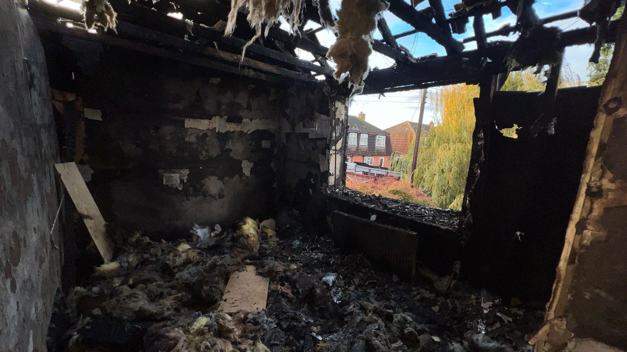 A bedroom on the upper floor of a house. The window is missing and fire has torn through the roof so that the sky is visible through the remaining rafters. The entire room is destroyed and black and insulation from the loft is littering the floor and hanging from the charred roof timbers.