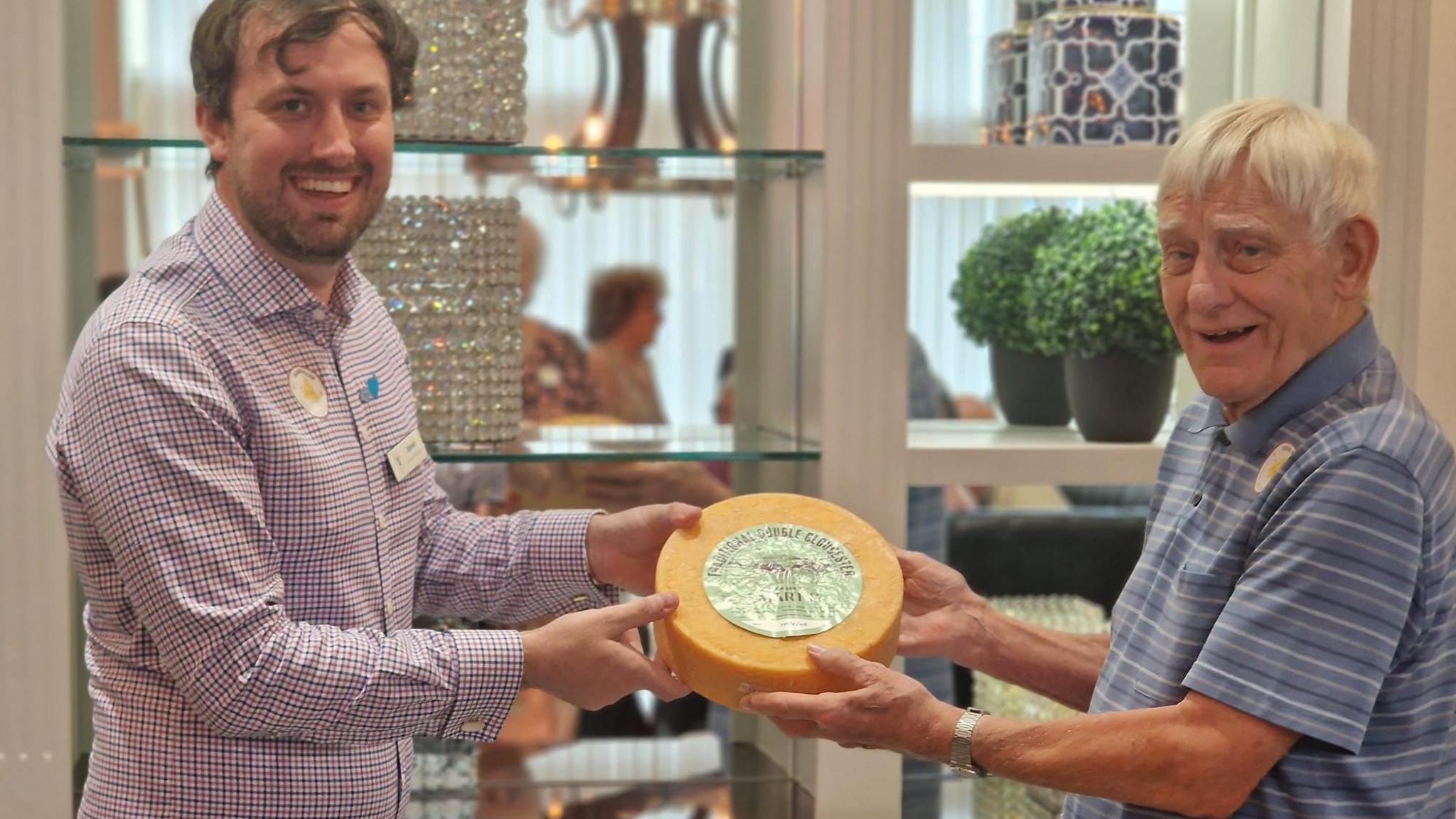 Two men holding a wheel of Double Gloucester cheese. The man on the right is elderly, has white short hair and is wearing a blue striped polo shirt. The man on the left is younger and has short dark hair, facial hair and is wearing a checked shirt. Both of them are looking at the camera and smiling.