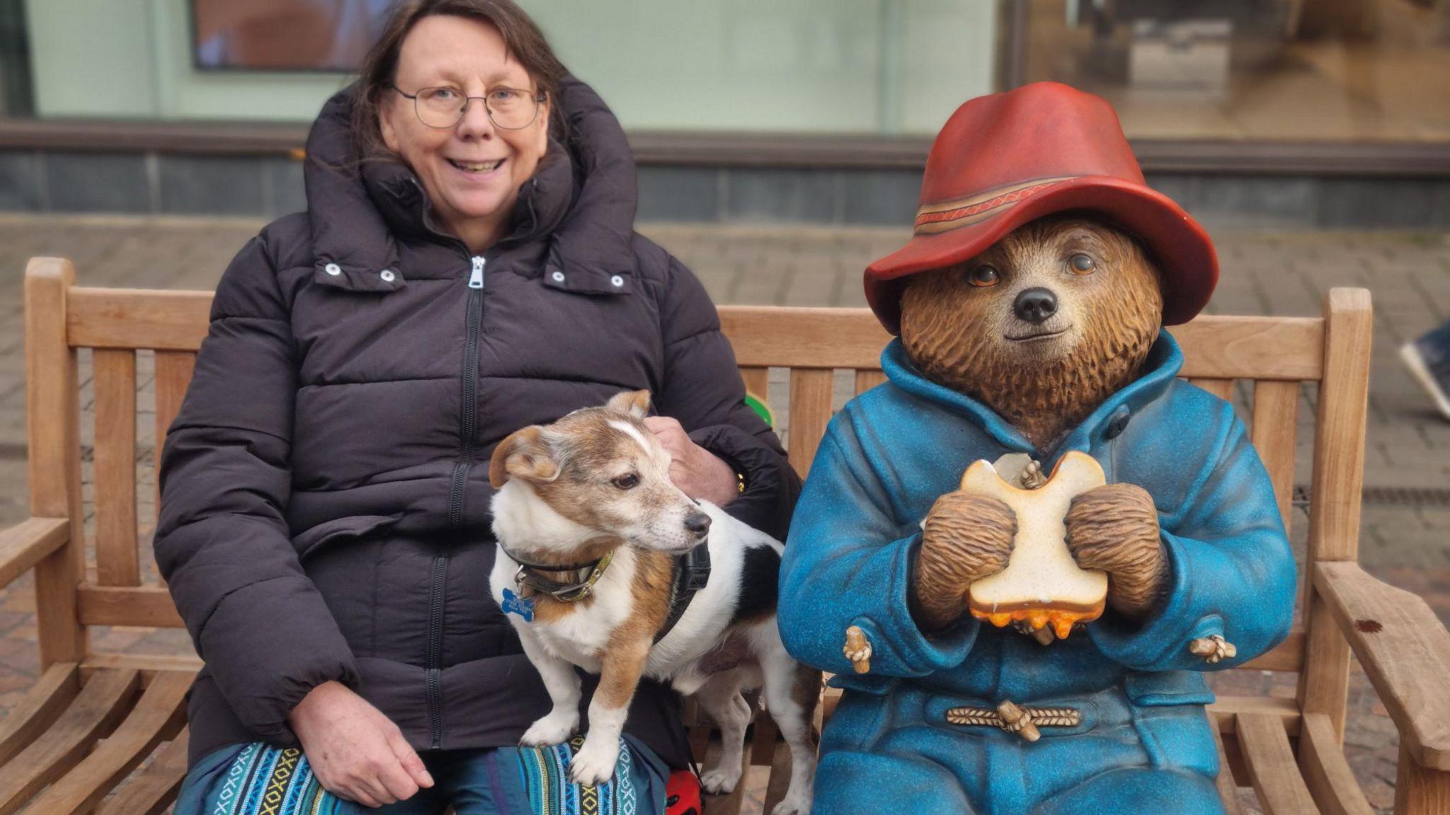 A woman smiling on the Paddington Bear bench. She has a dog with her. It is an overcast day.