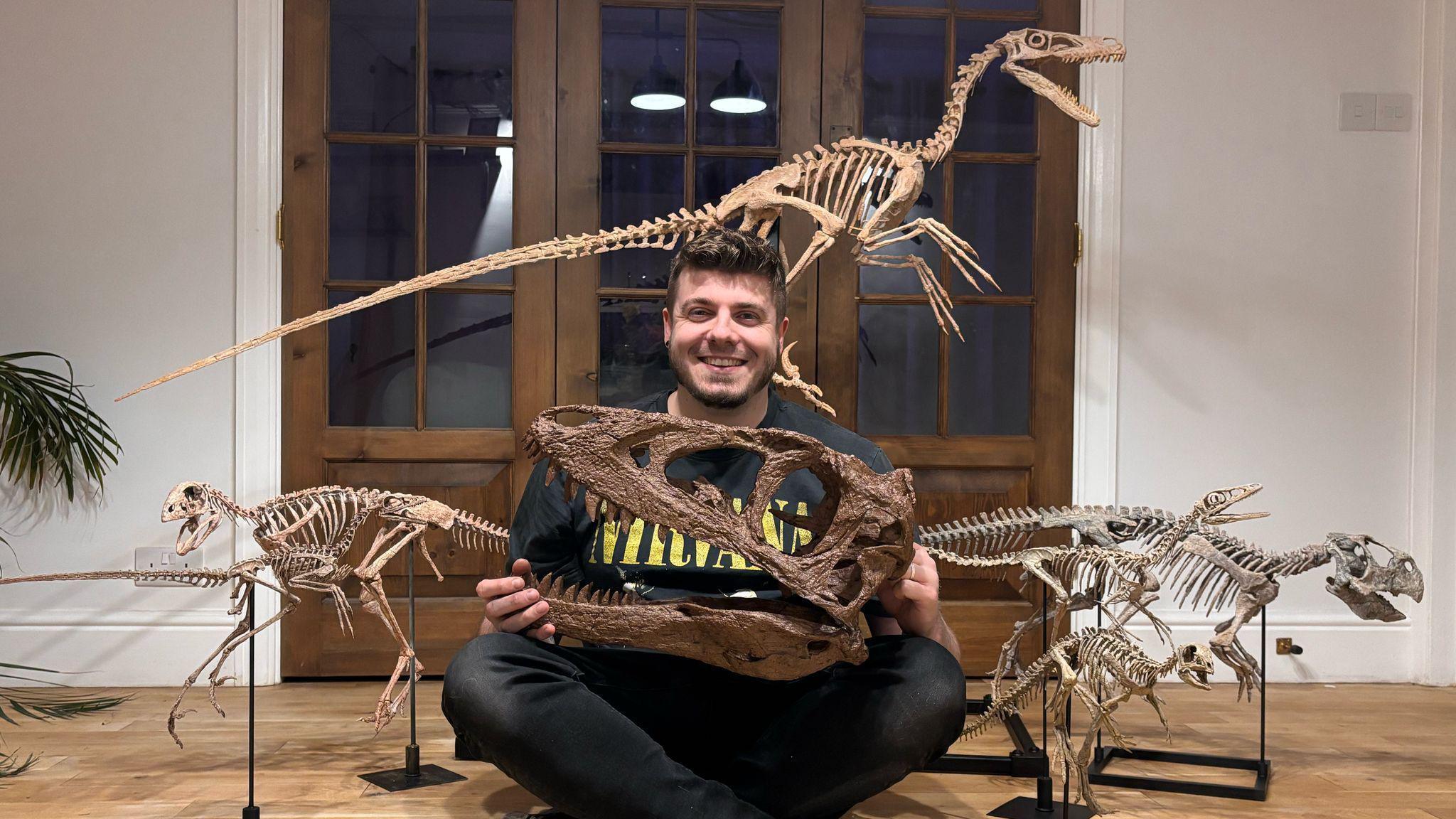 Mr Pritchard sits crossed leg on the floor, smiling at the camera, while holding the replica of a dinosaur skull. Small full replicas of other dinosaurs are placed either side of him and one behind him.