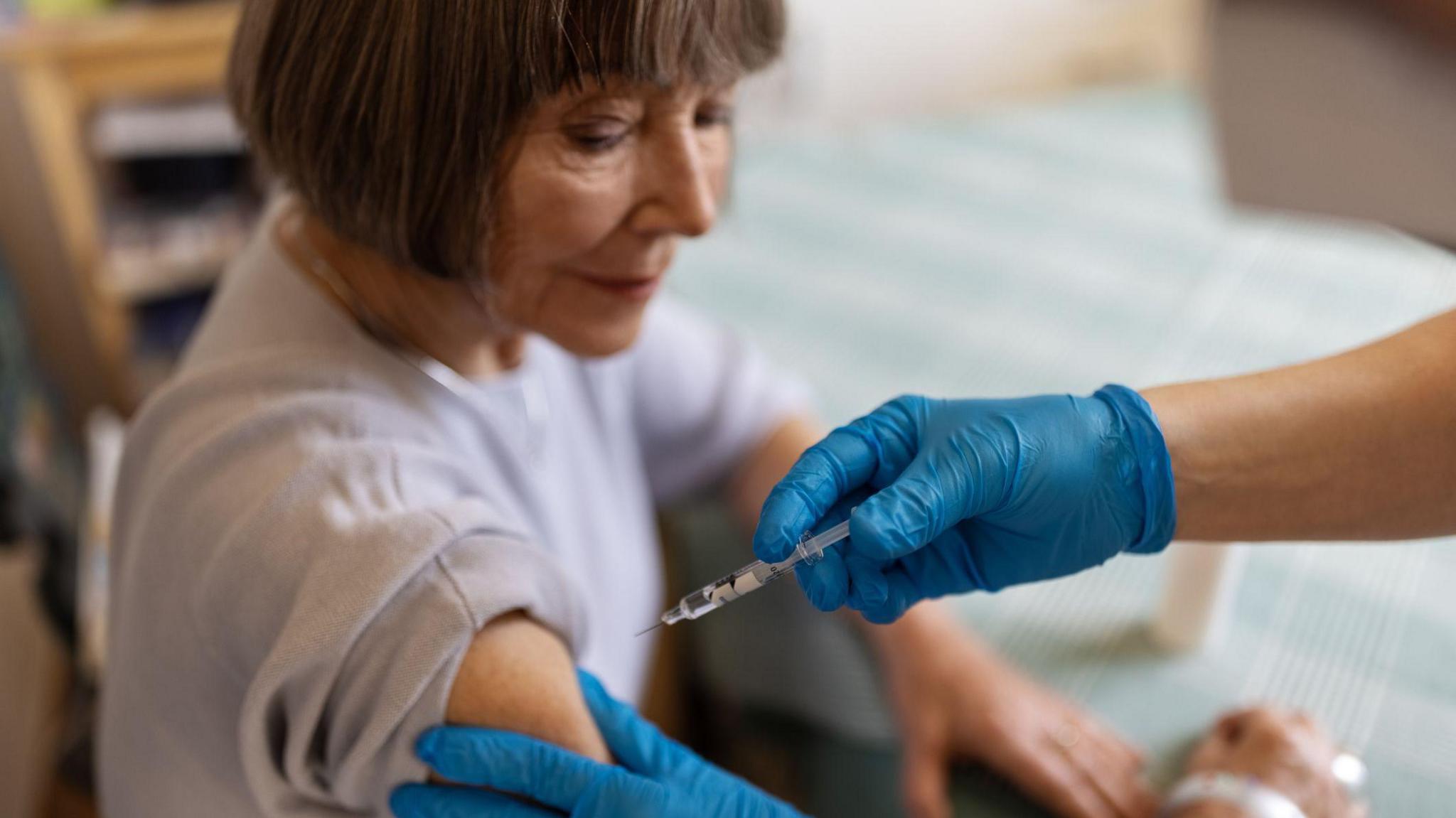 Generic picture of an older lady - with grey hair in a bob and a lilac t-shirt on - being given a jab by a medic in blue plastic gloves. 