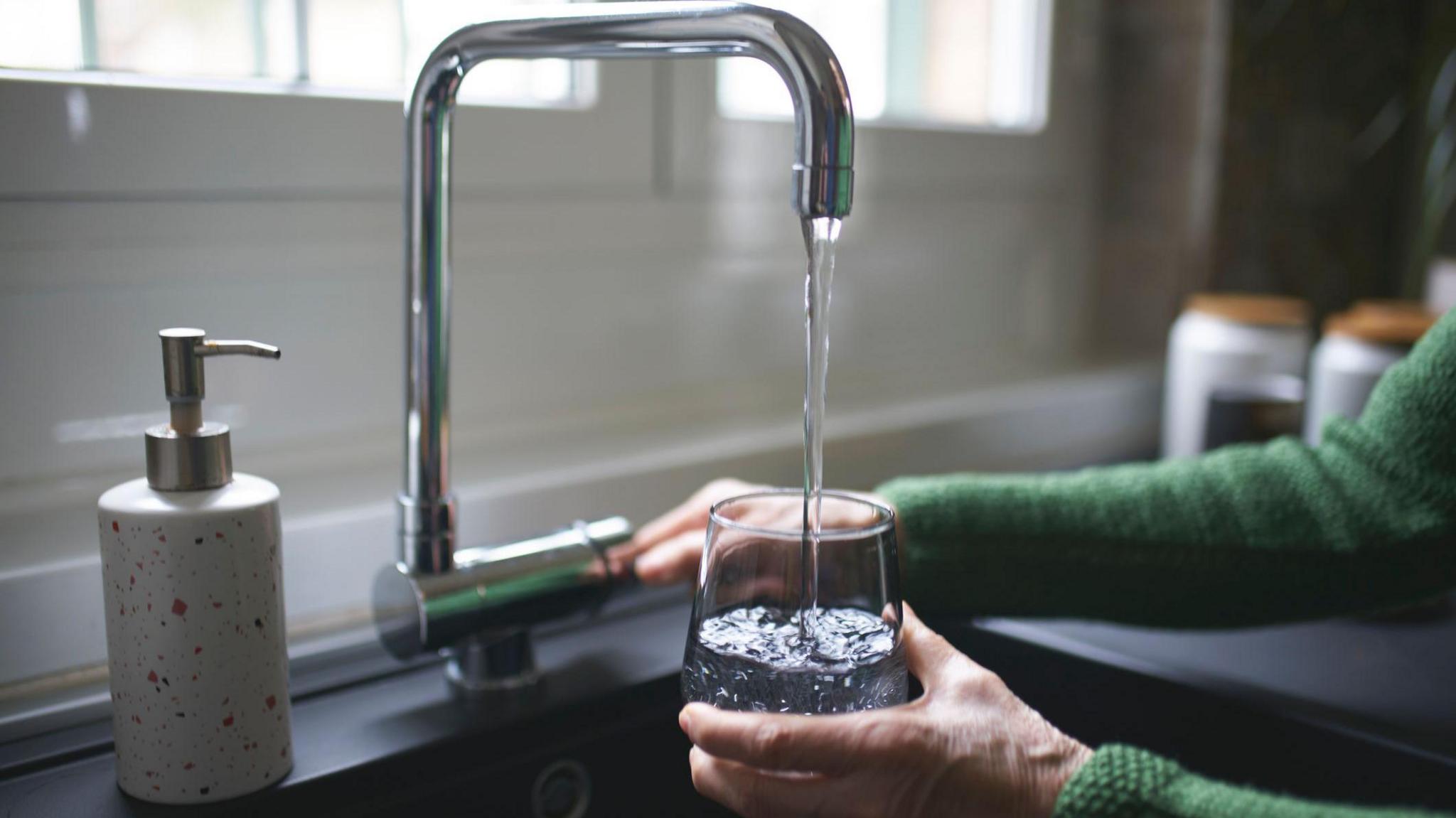A woman is filling a glass with water from a silver tap. You can only see her hands. She is wearing a green cardigan.