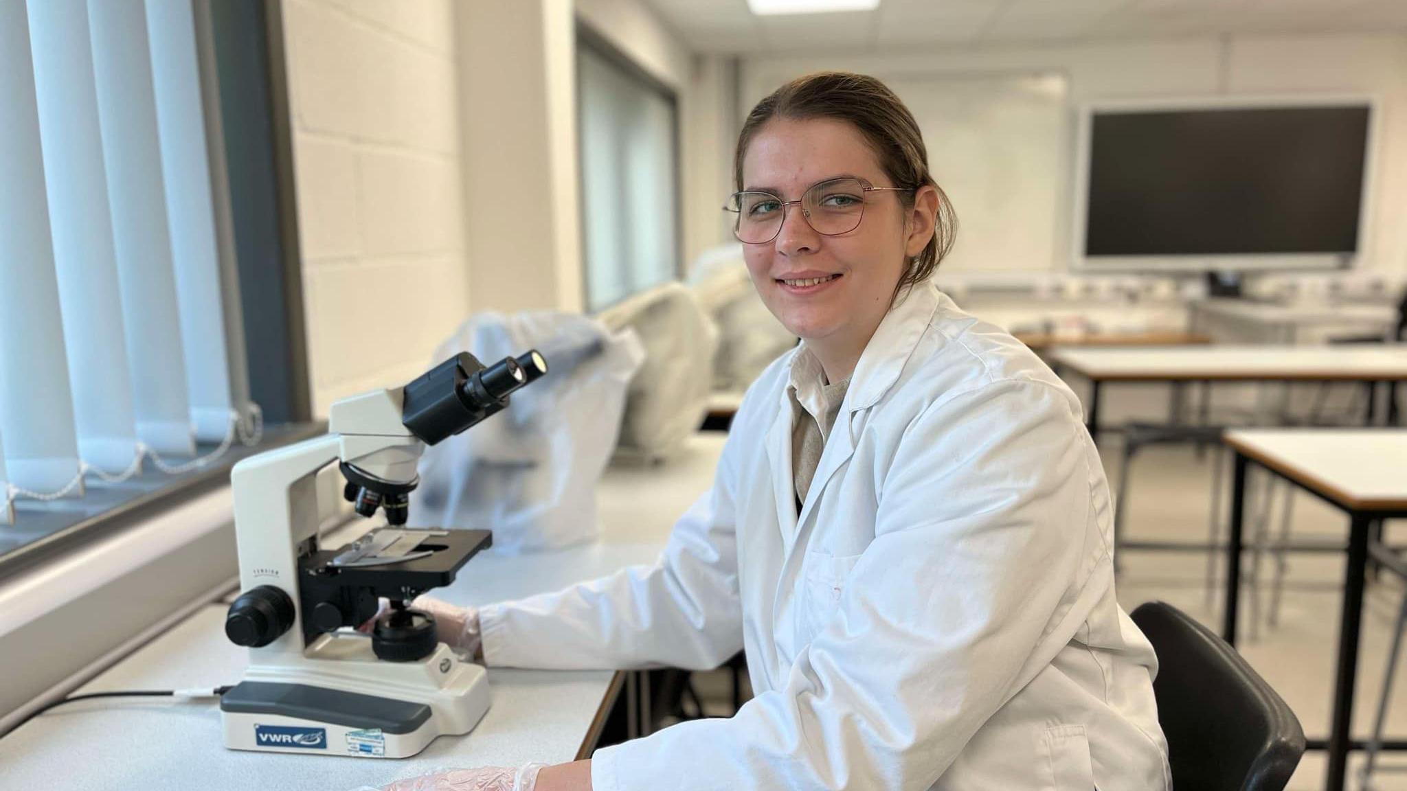 A woman wearing in lab coat and glasses, sat at a desk with a microscope. Other tables can be seen in the background in a classroom.