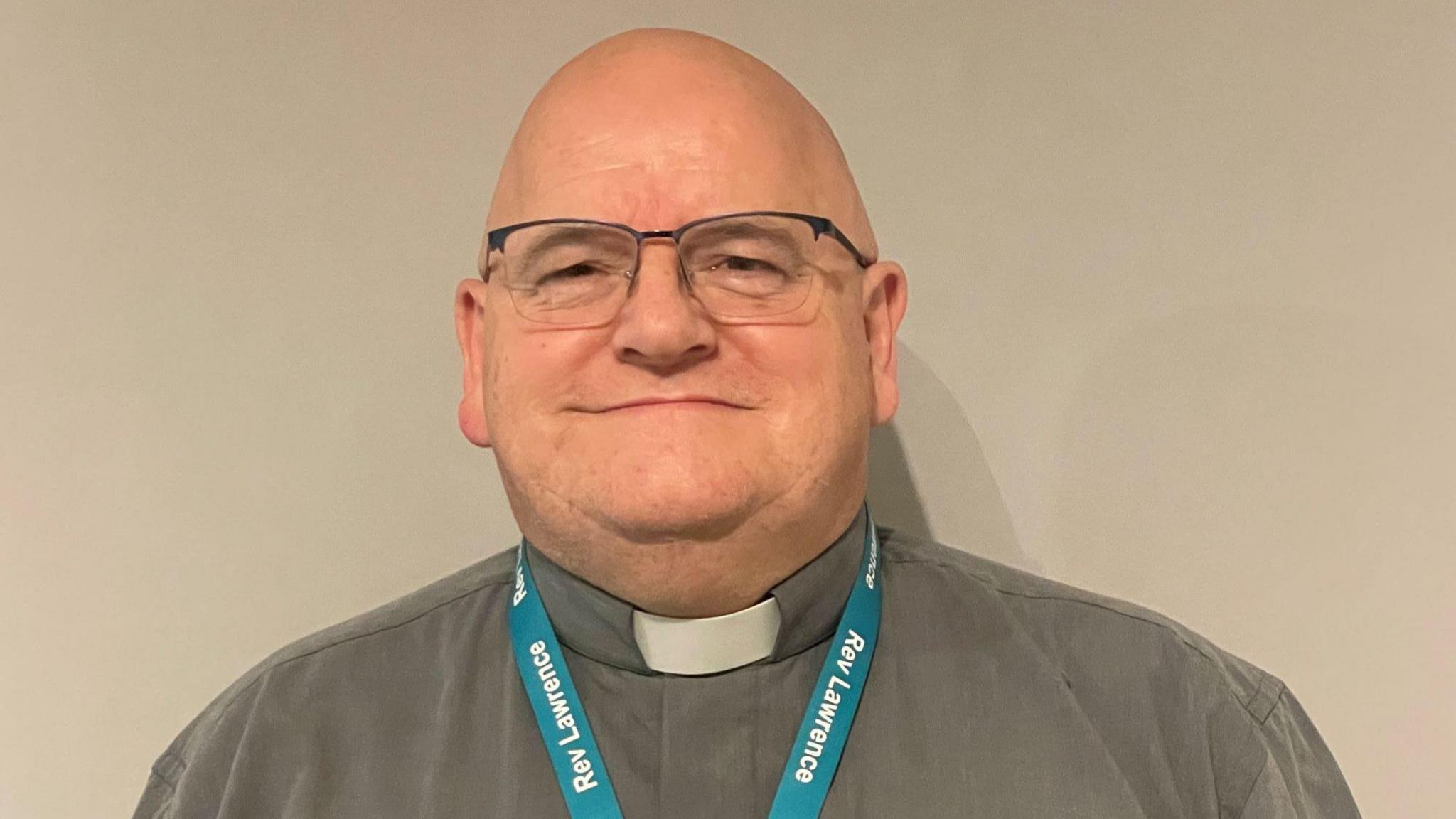 Lawrence Carey smiles at the camera. He is wearing a grey shirt with a white dog collar. He has glasses and is wearing a lanyard with his name on. 