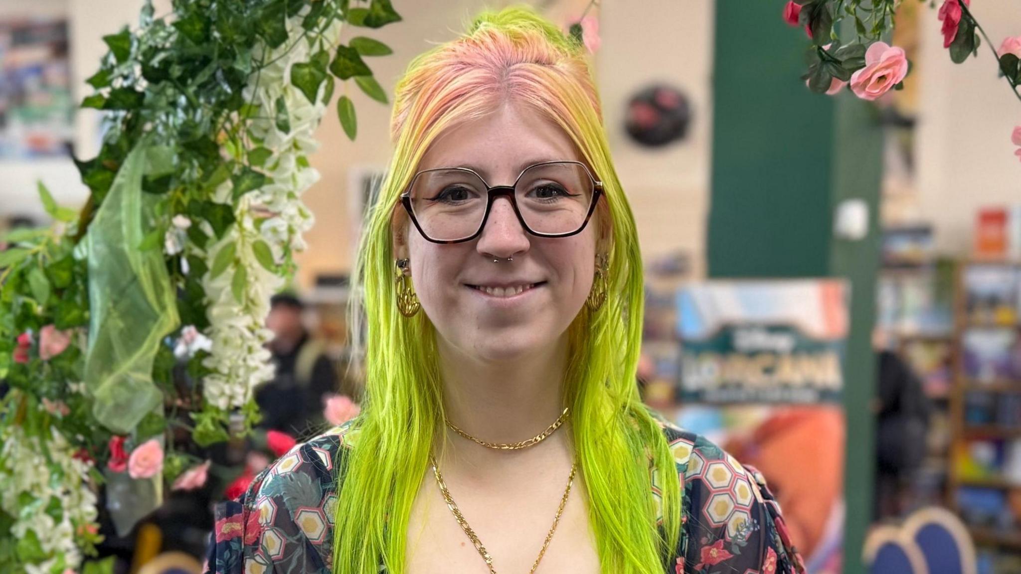 Amy Wolsey a young white woman with bright green hair with pink roots. She is wearing a floral dress with a gold necklace. She is standing in a game board café which she manages.