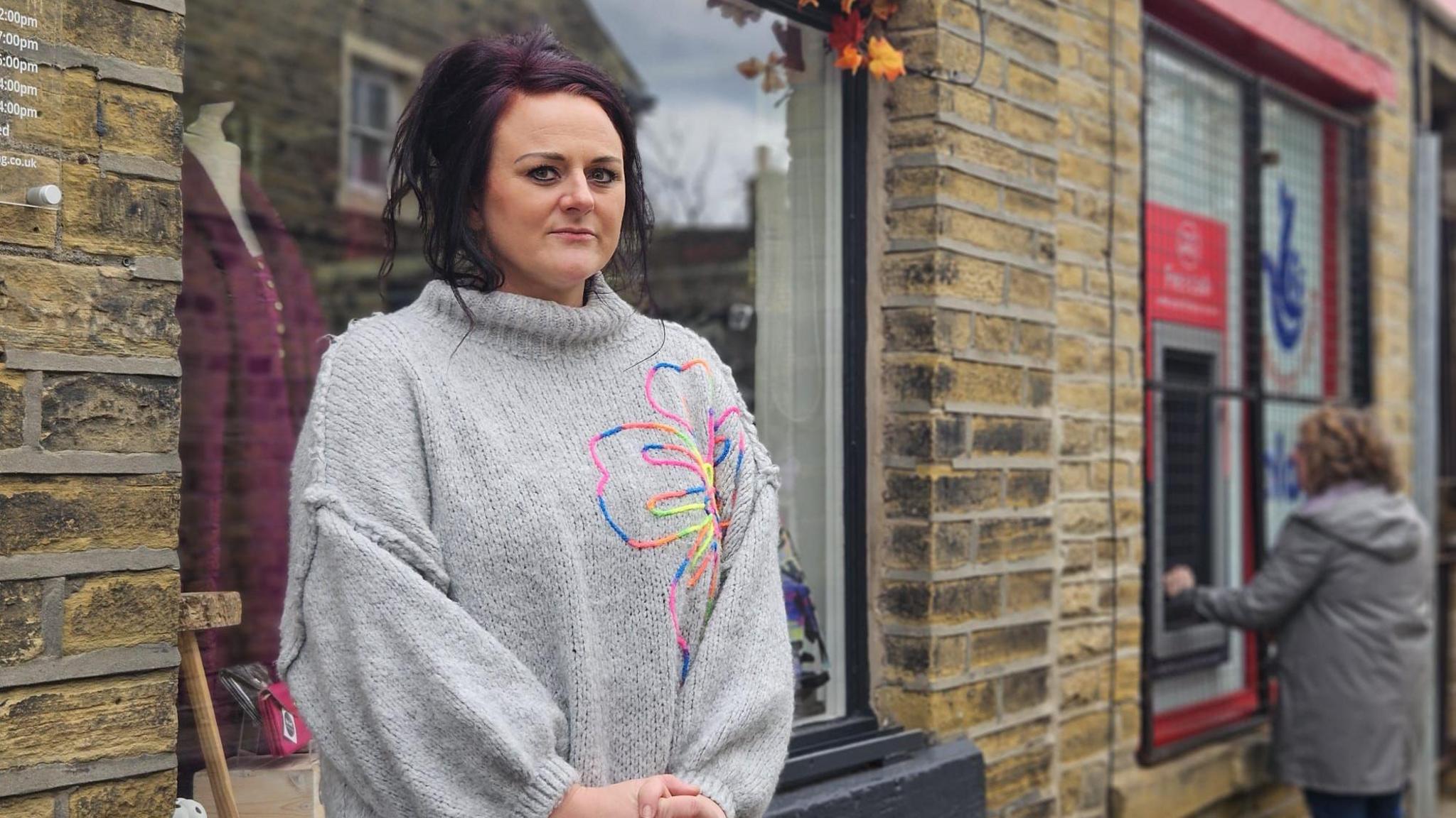 A woman stands outside a shop front looking serious. She wears a grey jumper with a large floral pattern on it. In the background is a woman outside a post office, using a cash machine.