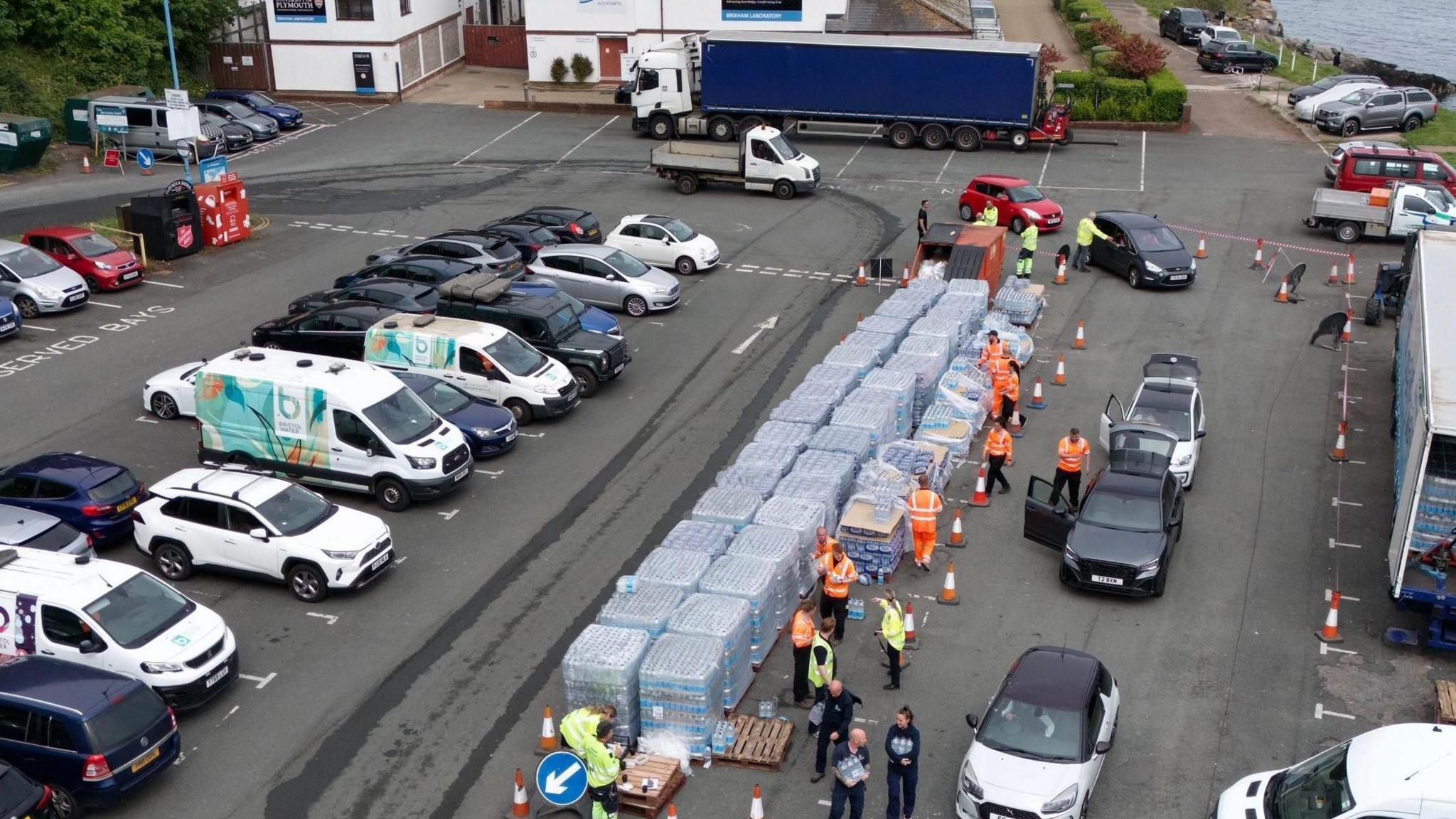 An aerial view of a carpark where stacks of bottled water are being collected by people and taken to their vehicles. 