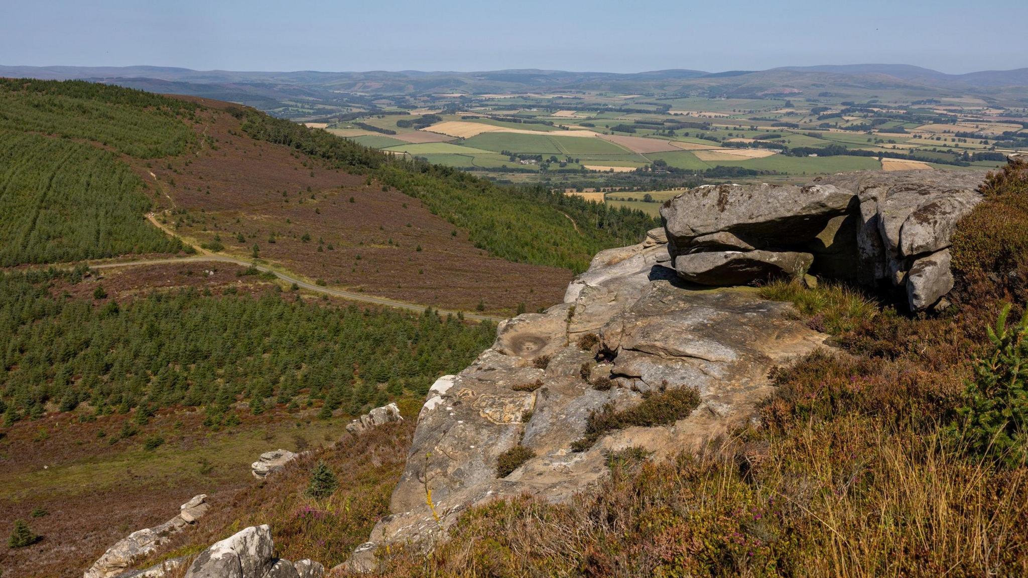 A view of the Rothbury Estate which has rocky outcrops overlooking a vast expanse of land. Below there is lots of green, brown and yellow fields and farmland with hills visible in the distance.