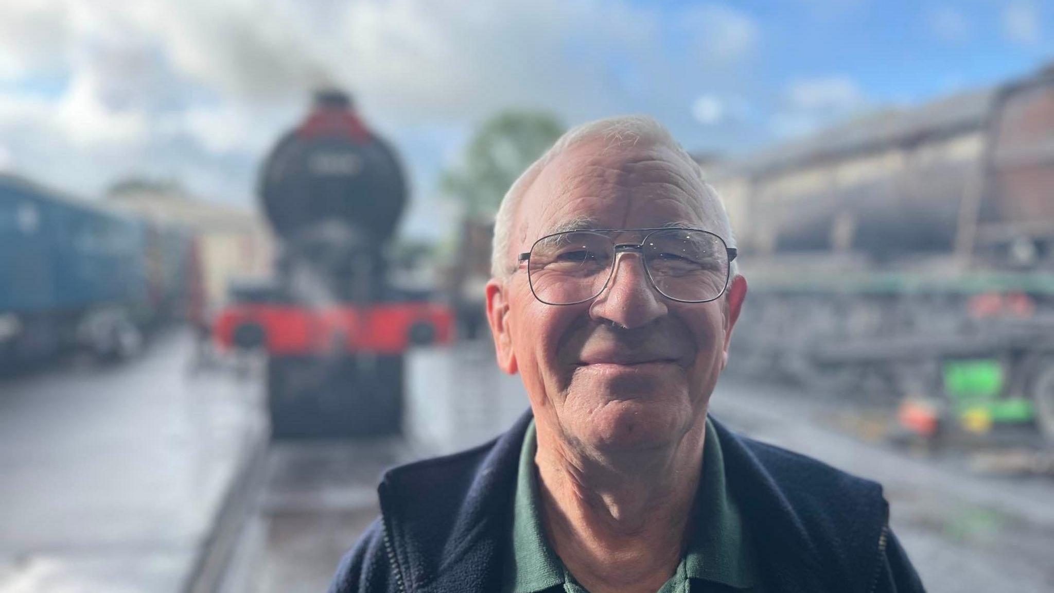 An man with grey hair and large glasses smiles at the camera. 