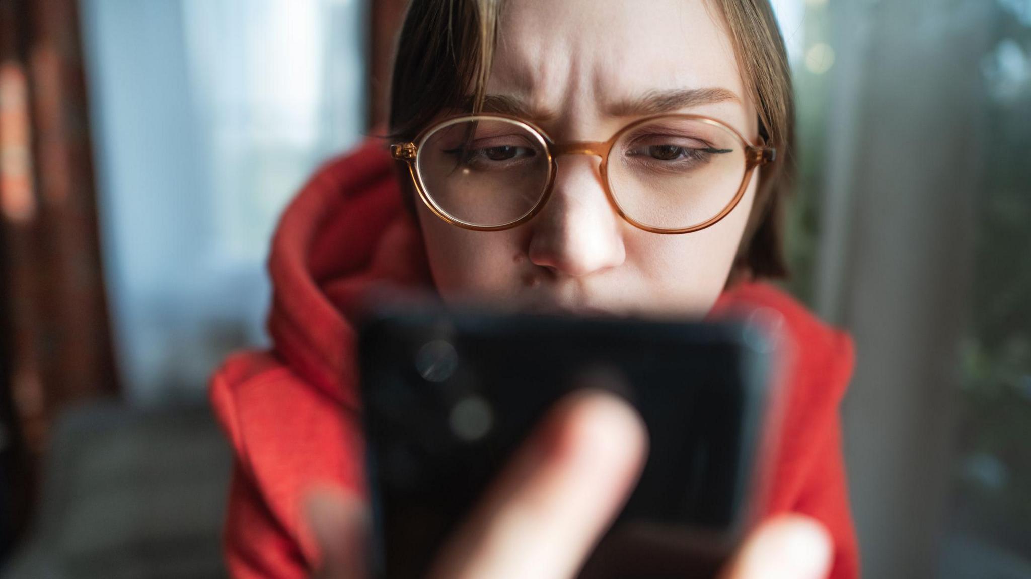 Woman wearing glasses looks at phone which is prominent in the foreground.