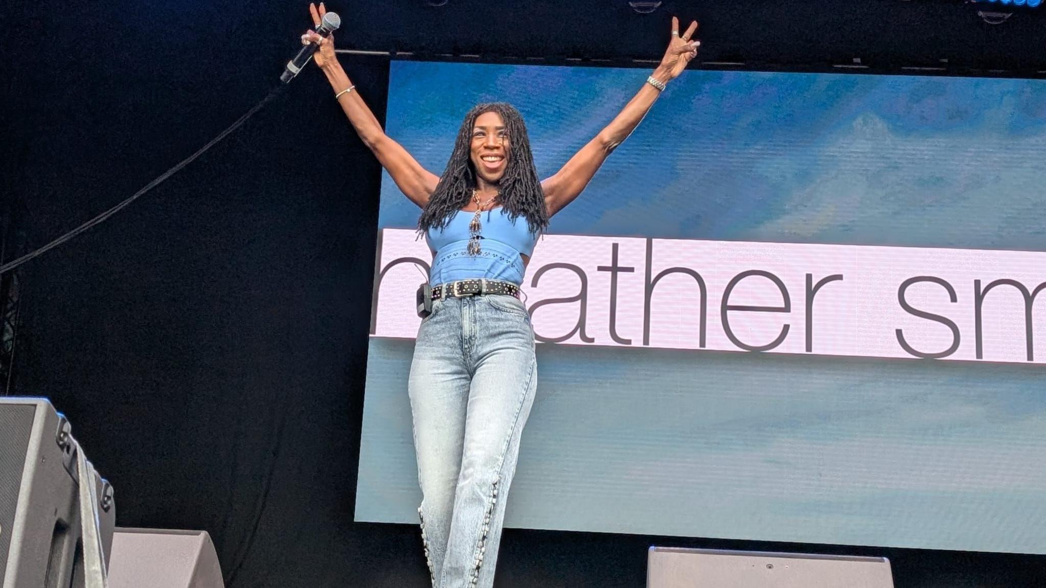 A woman stands on a stage and holds up a microphone, while she also does the peace sign with both hands. She has long black braids, and she is wearing jeans and a blue vest.