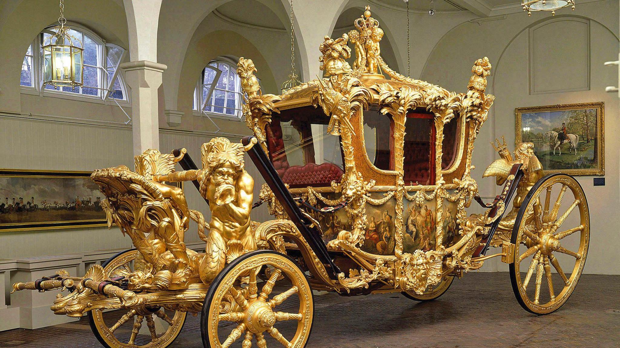 A huge ornate gold covered coach in the Mews. It has painted doors and panels and decorative figures. 