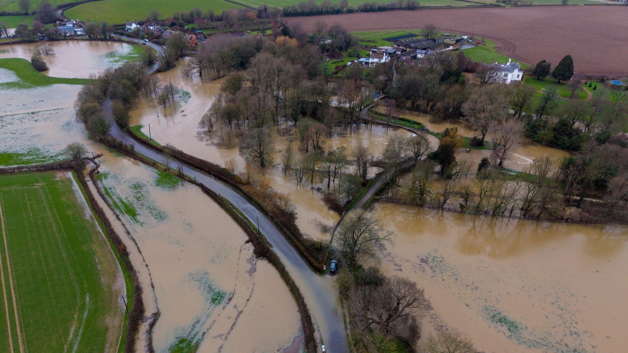 A curving country road through fields and a small collection of homes, seen from above. The fields and part of the road are flooded with brown water.
