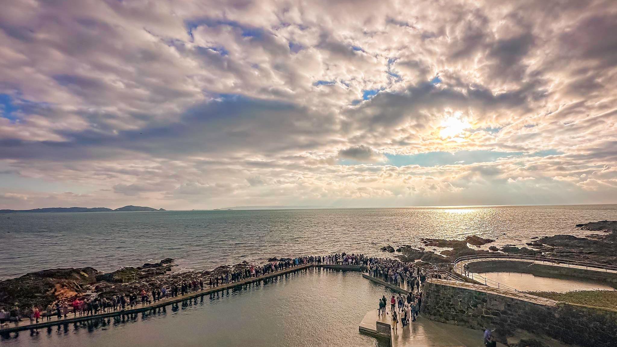 Hundreds of people gathered at La Vallette bathing pools on the seafront in Guernsey. It is a sunny day and the sea is relatively calm.