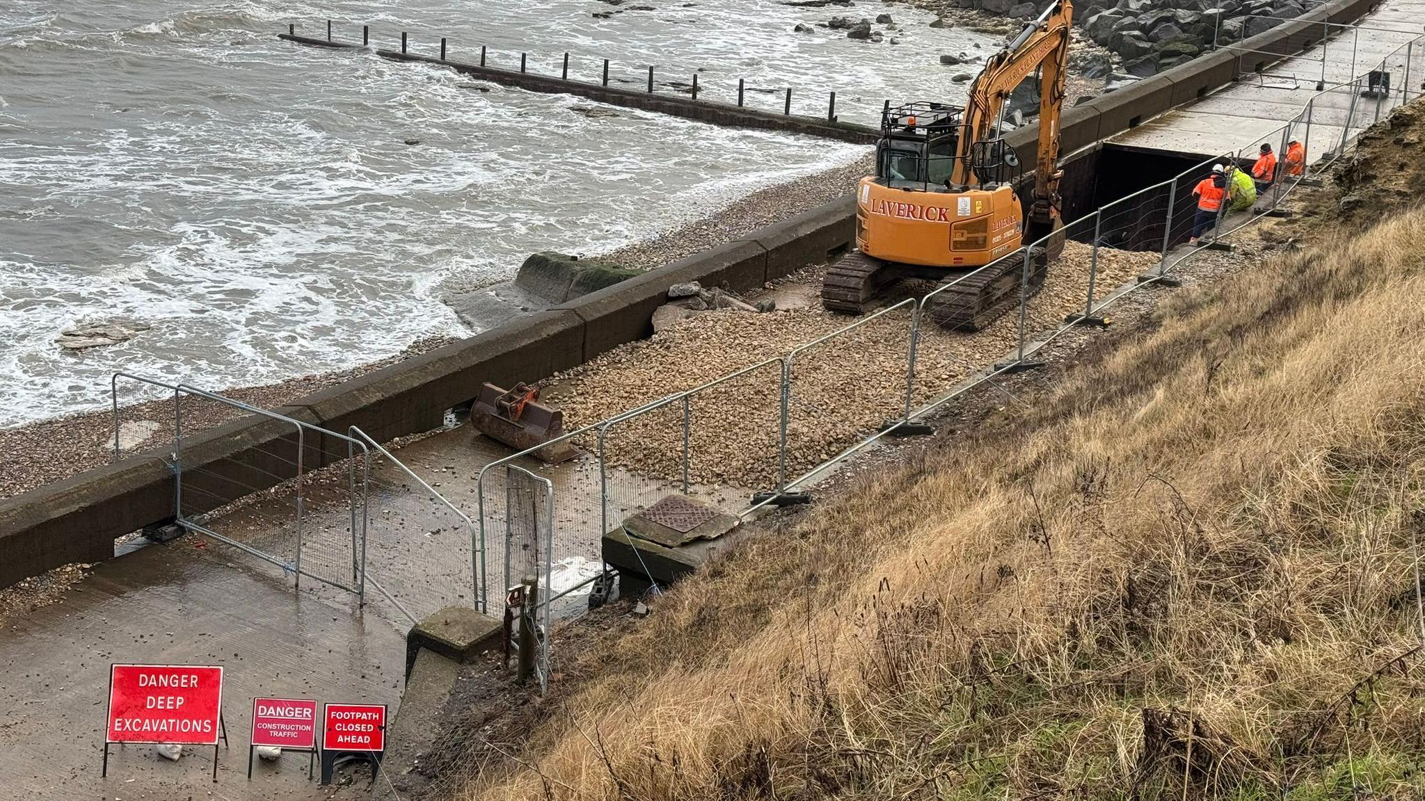 A yellow digger carrying out repairs on the collapsed section of the promenade.  Four workers in high visibility vests are looking on. The area is fenced off with red signs warning about danger are around it. 