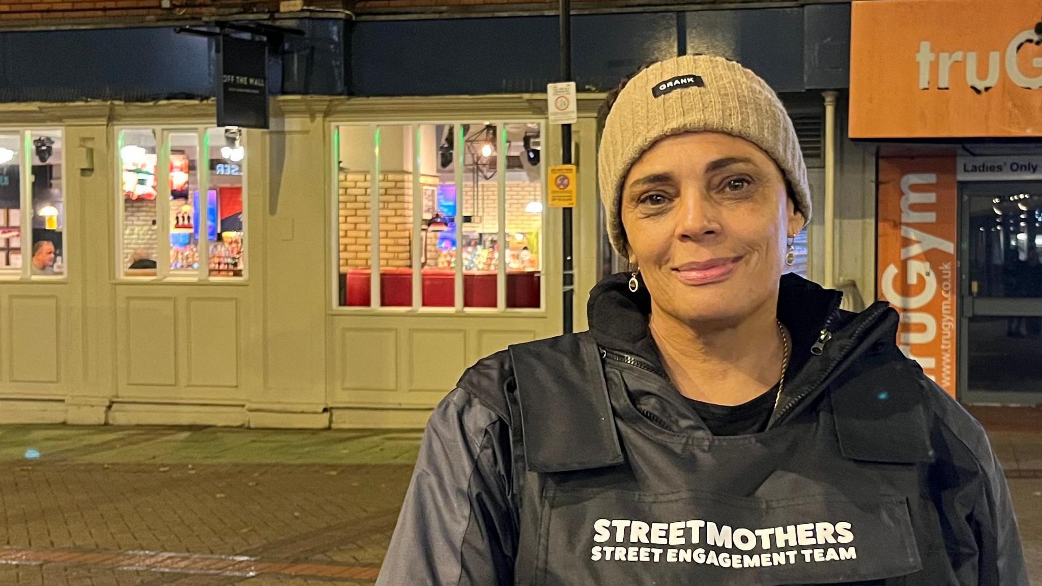 Sharon McGovern is standing on a street at night wearing a black stab-proof vest with white text that reads: “STREET MOTHERS STREET ENGAGEMENT TEAM.” Ms McGovern also has a light-coloured cap on. Behind her is a row of brightly lit shops.