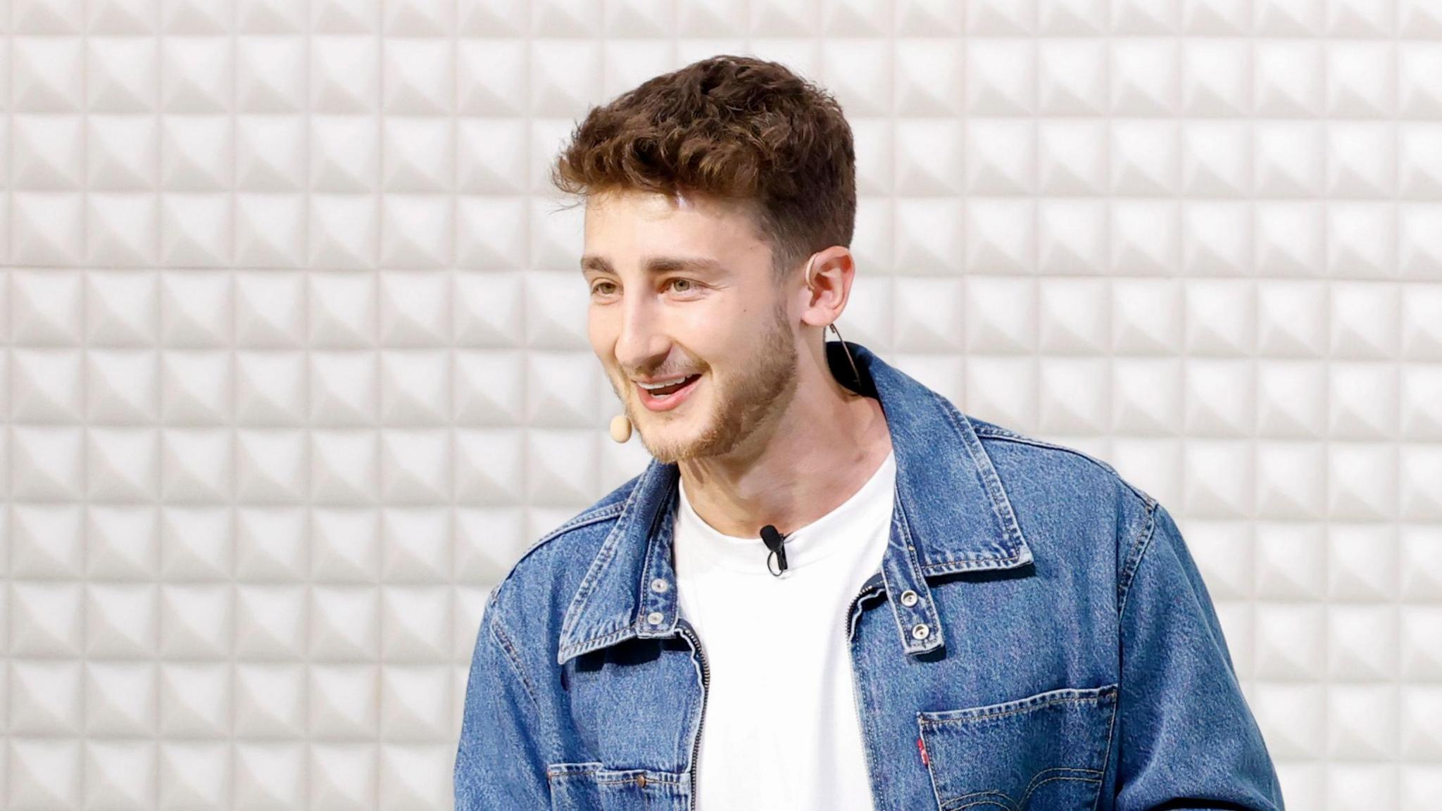 Brandon B wears a jean jacket and white shirt. He is presenting at an event, with a microphone attached to his shirt and also on his cheek. Behind him is a simple padded white background.