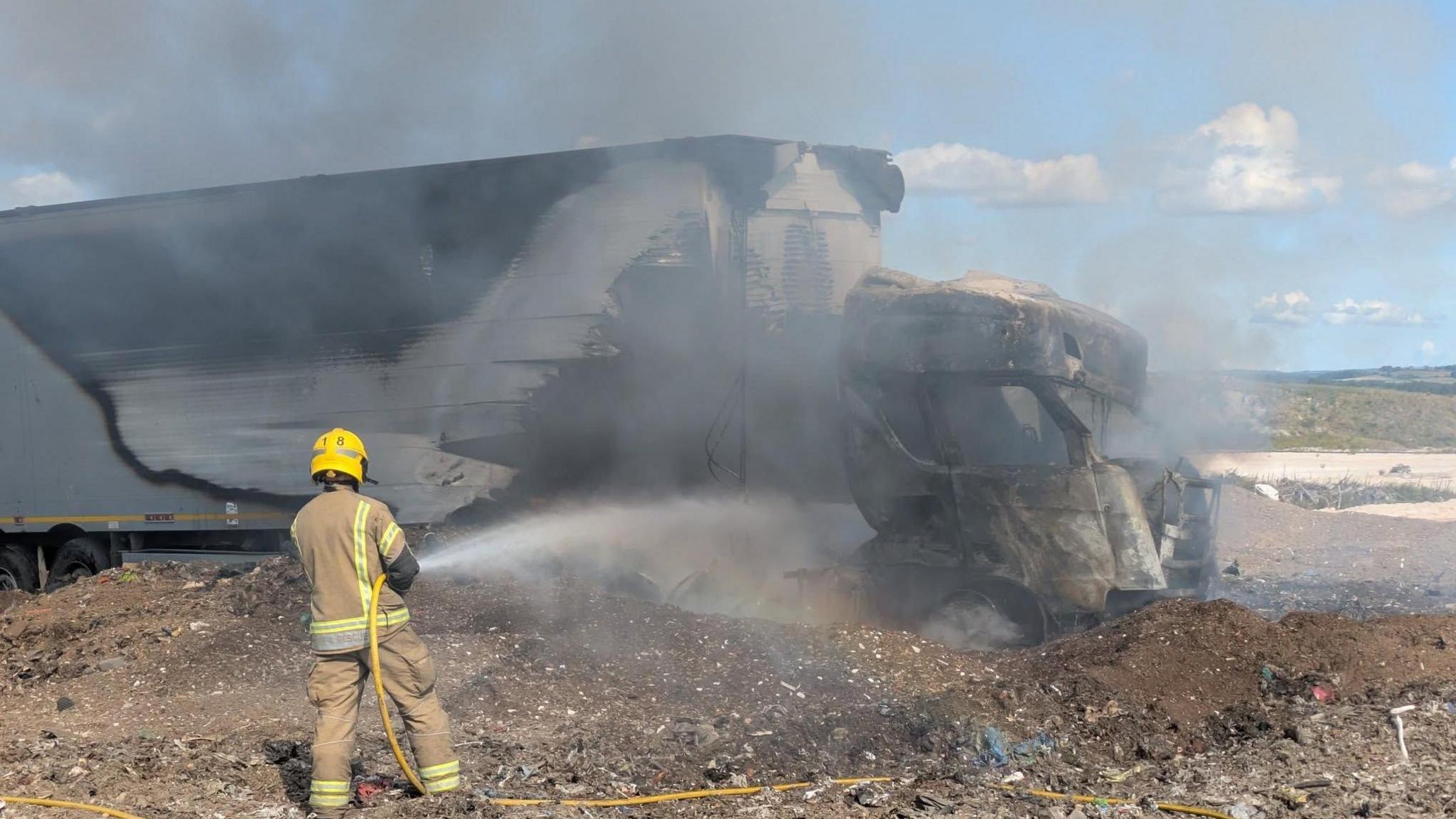 A firefighter with hose spraying water onto a burning lorry surrounded by rubbish.