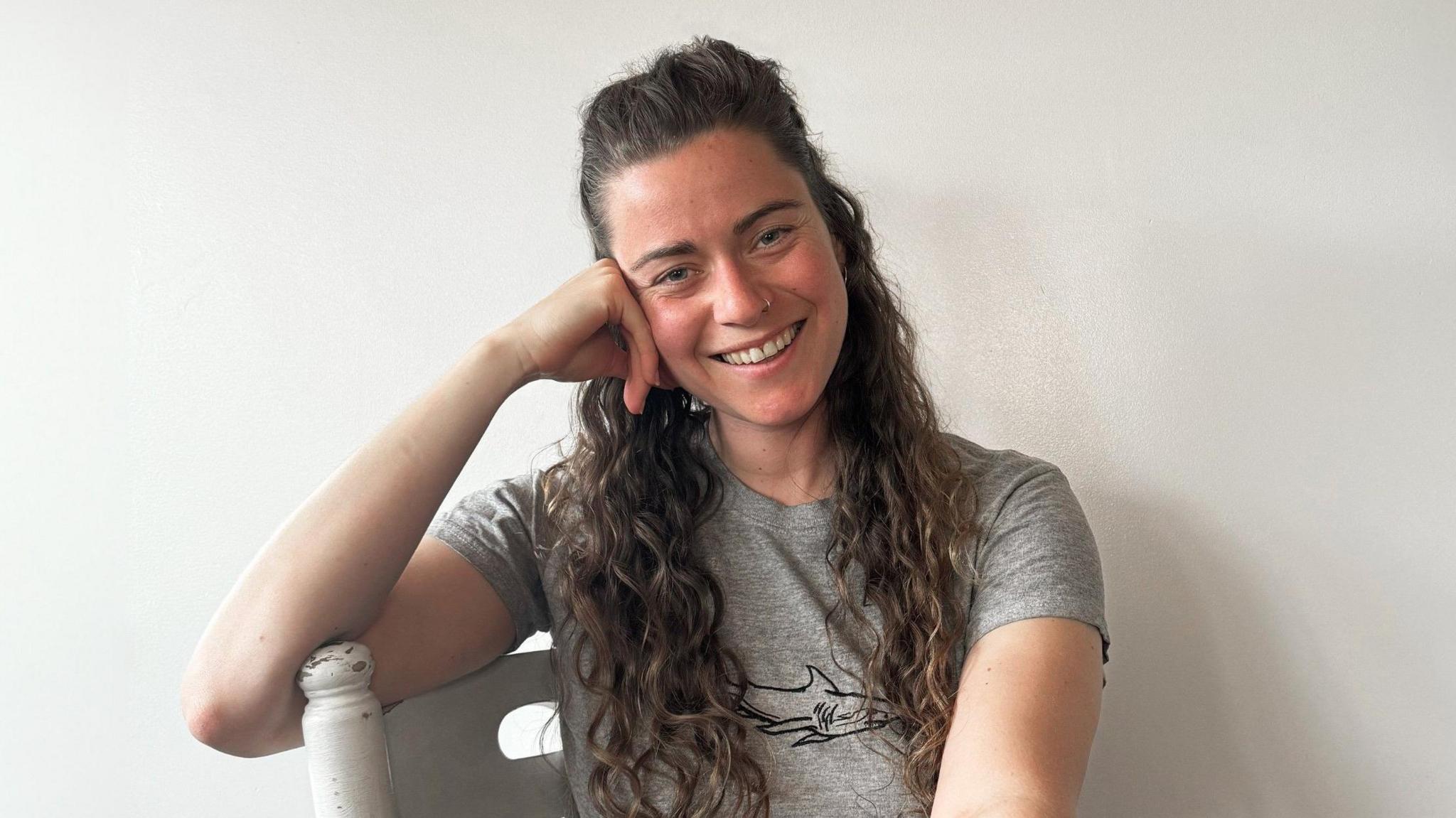 Charlie Gill rests her head on her hand while leaning on a white painted chair. She wears a grey T-shirt with a shark printed on it and smiles at the camera. She has long curly brown hair which she wears half-up-half-down