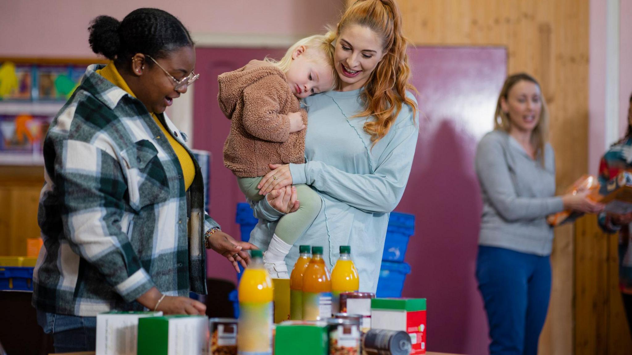 A woman with long ginger hair tied back in a pony tail and wearing a long-sleeve grey top is collecting groceries and toiletries from a woman who is standing next to a table full of food and drink items.