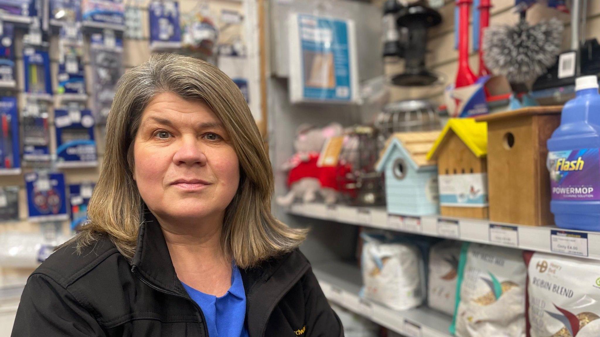 Anne is looking at the camera. She has shoulder length hair and a black jacket on. In the background are products in her shop such as bird feeders and mops and buckets.