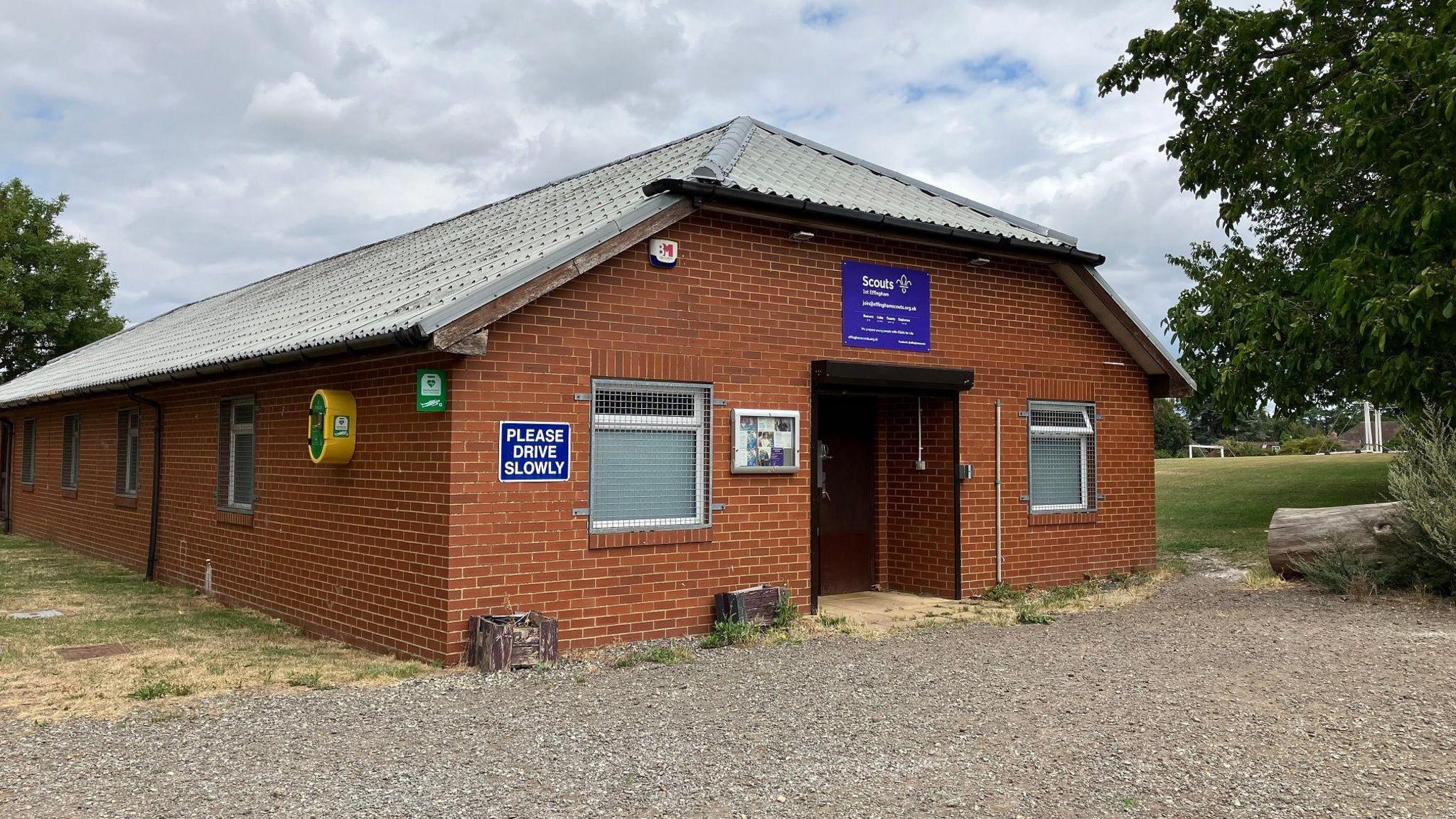 A low brick building with a pointed roof. There is a please drive slowly sign and a scouts sign above the door. Playing fields can be seen in the background