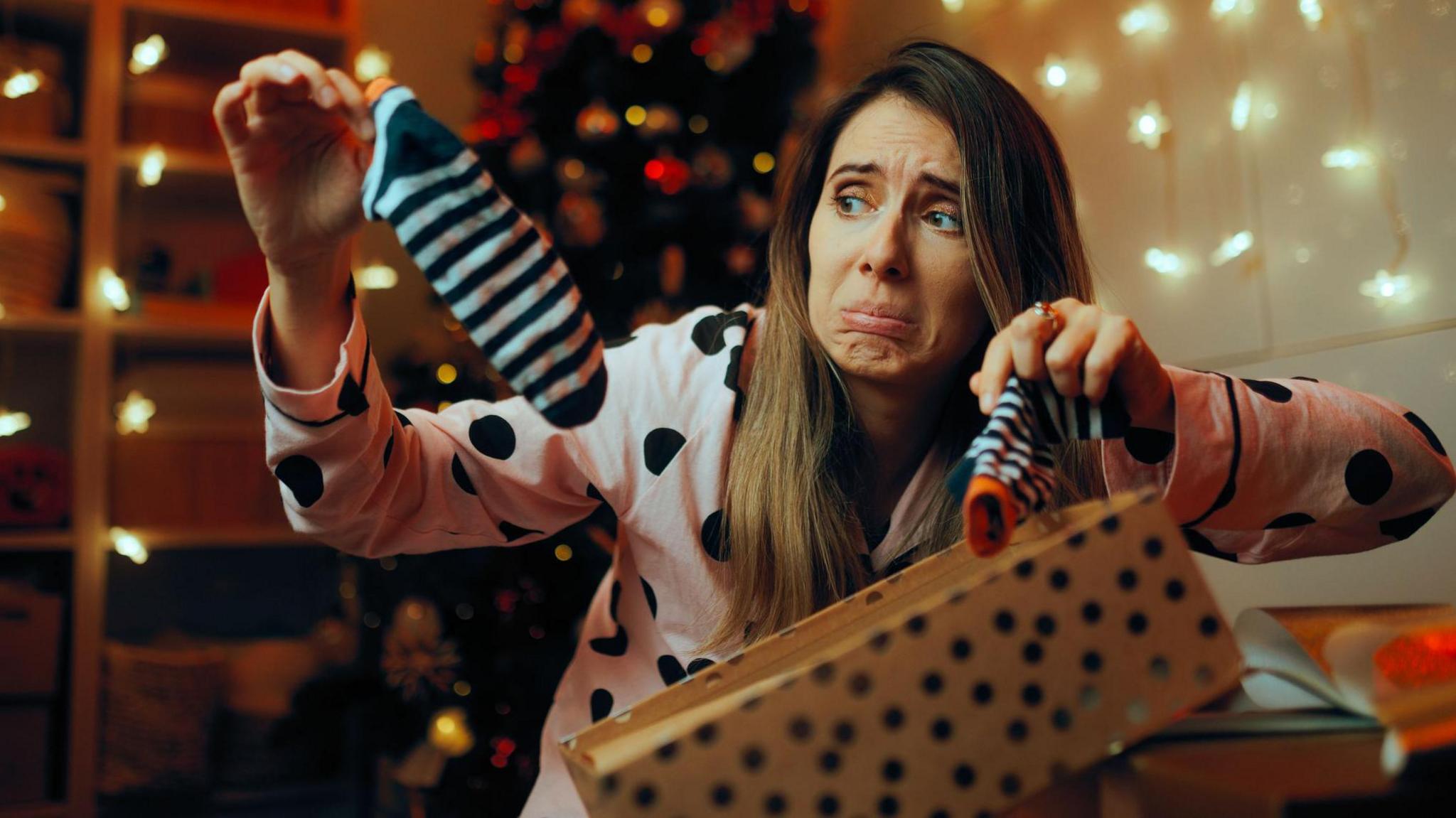 A woman in a pink long-sleeved shirt with black spots opens a gift and holds up two black and white stripy socks from it. She looks unhappy at the socks. She has long brown hair and brown eyes.