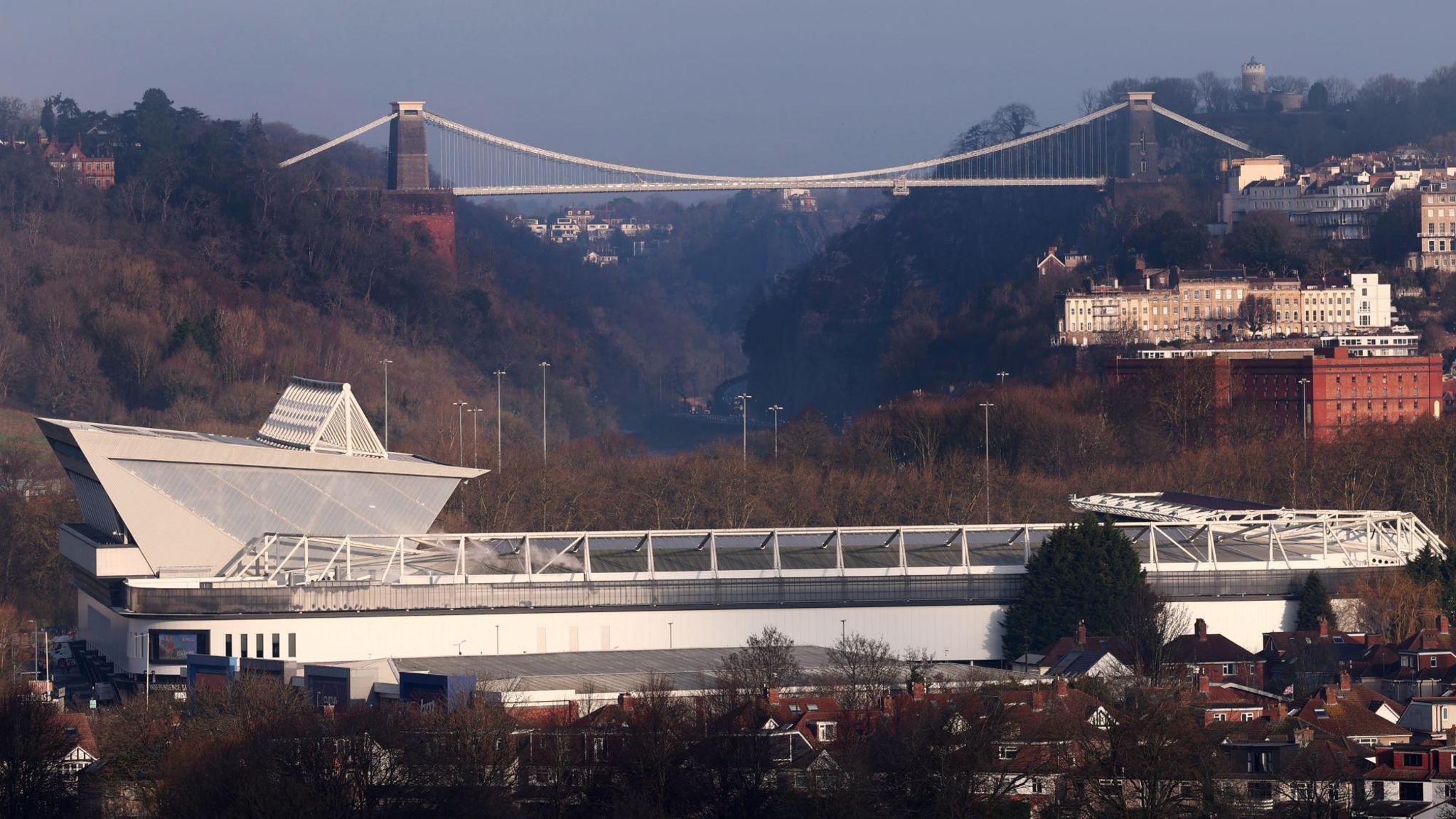 An aerial view across Bristol towards Avon gorge. Ashton Gate Stadium is in the foreground, surrounded by residential homes. In the distance, the Clifton Suspension Bridge hangs above the gorge, surrounded by trees. 